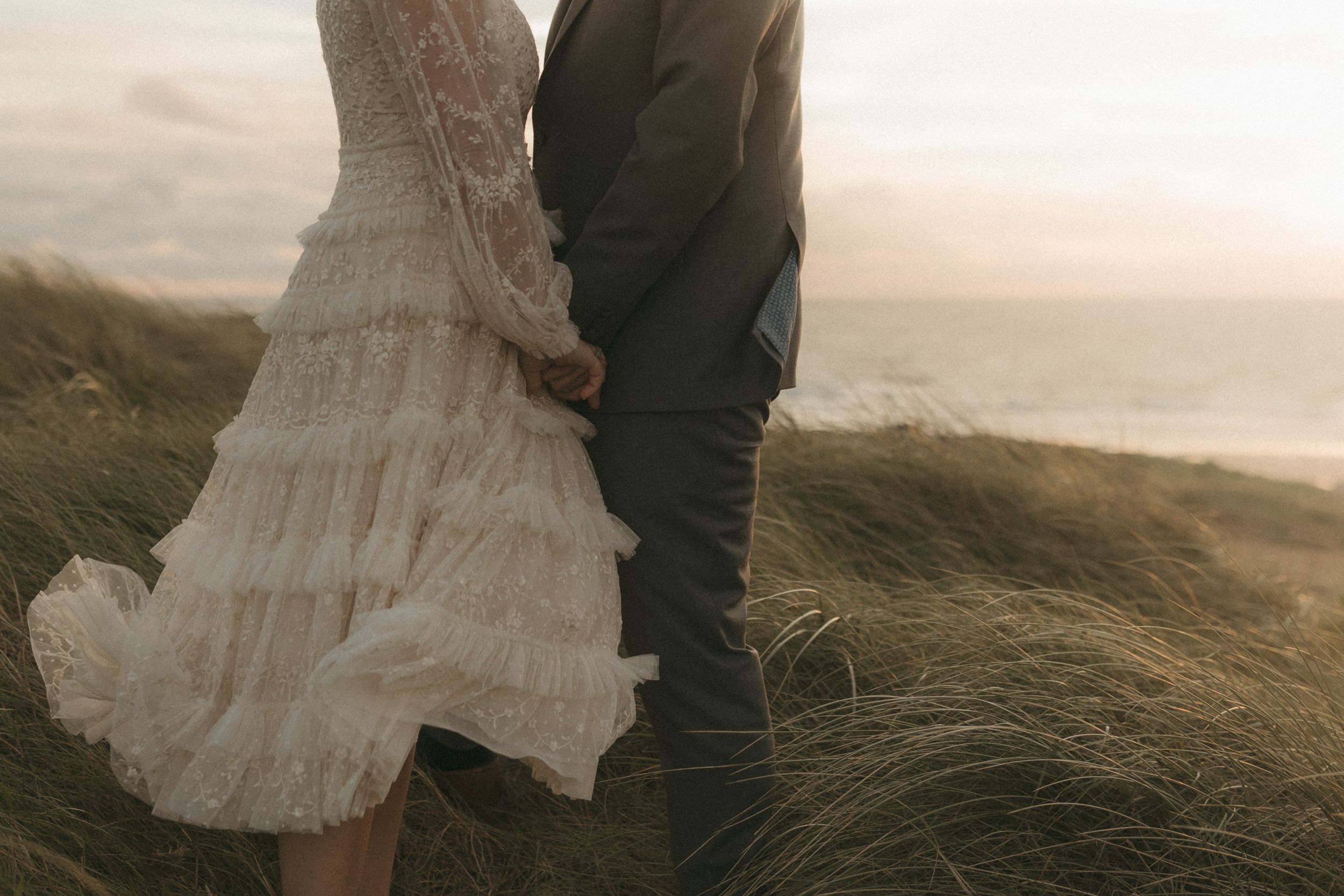 A couple in wedding attire walks through windswept coastal grass at sunset near rugged cliffs.