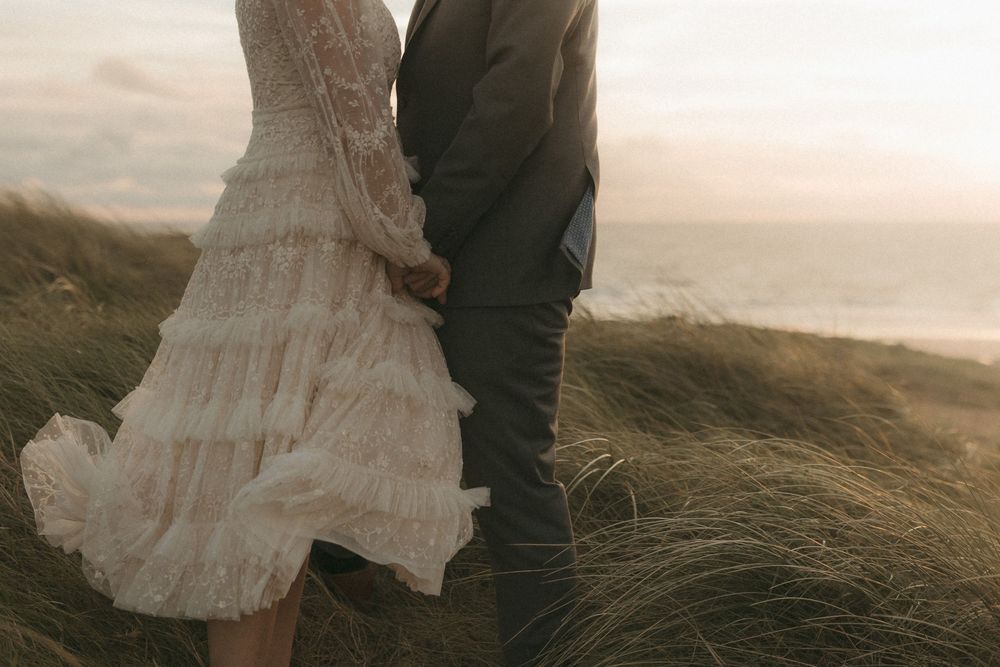 A couple in wedding attire walks through windswept coastal grass at sunset near rugged cliffs.