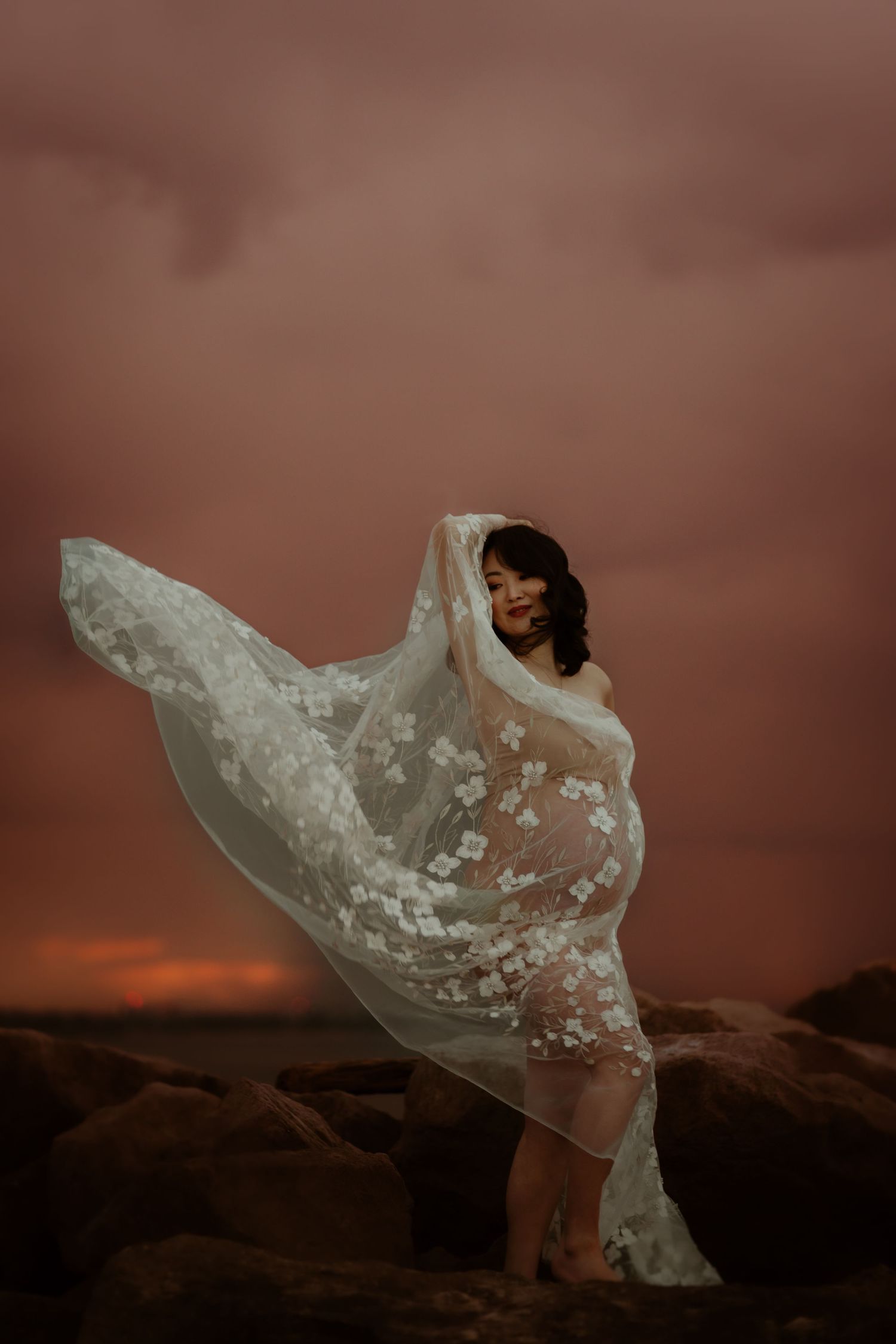 A white flowing dress catches the light of a dramatic sunset on a sandy path through coastal vegetation.