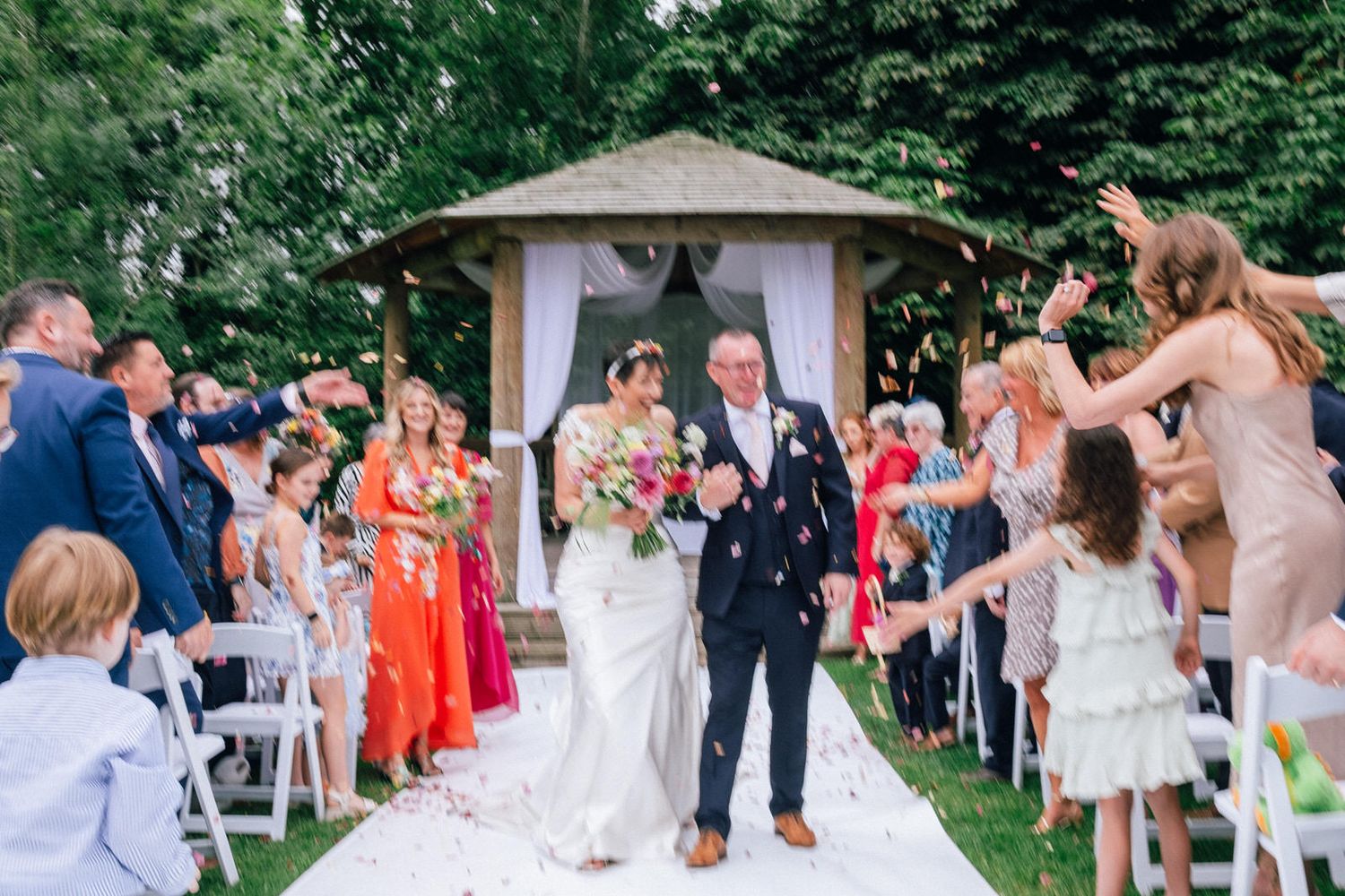 Wedding guests celebrate during an outdoor garden reception with greenery and decorative lighting.
