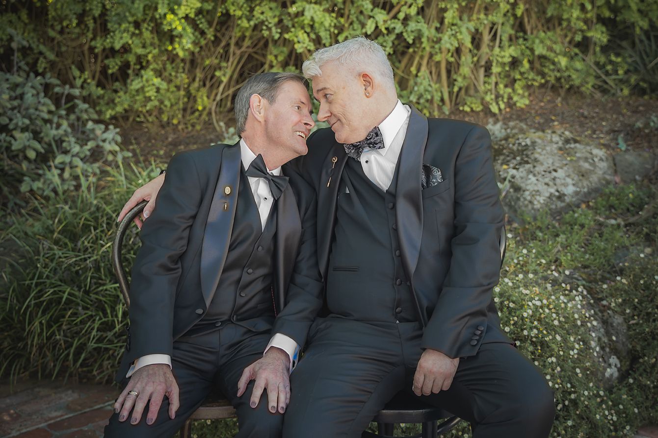 A couple in dark suits share intimate moments together in a garden setting during a wedding photoshoot.