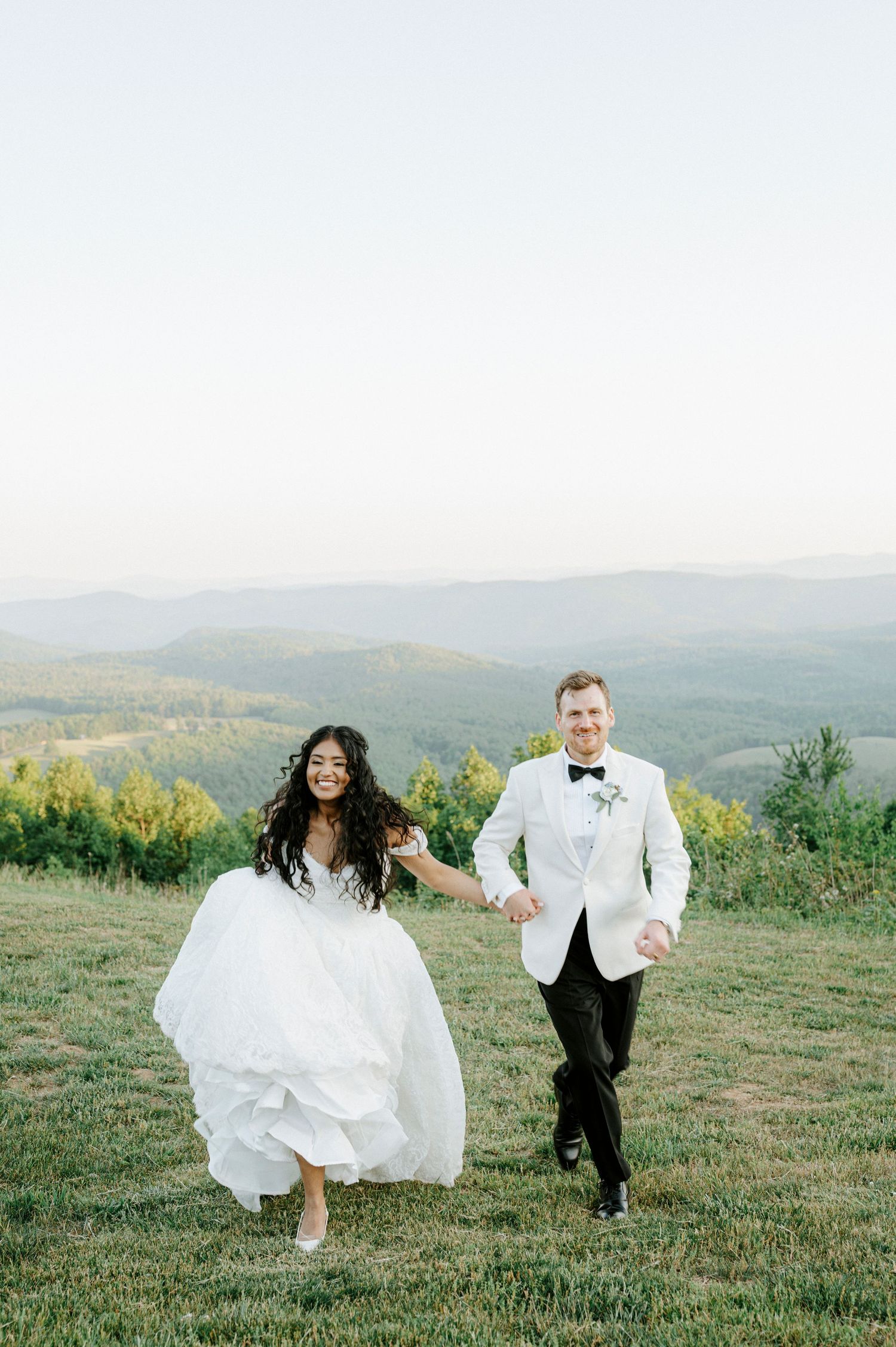 A joyful bride in a white gown and groom in a white tuxedo hold hands running on a hilltop with mountain views.