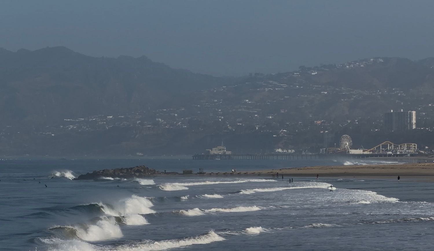 Coastal beach scene with rolling waves, misty mountains, and a seaside town visible in the background on a hazy day.