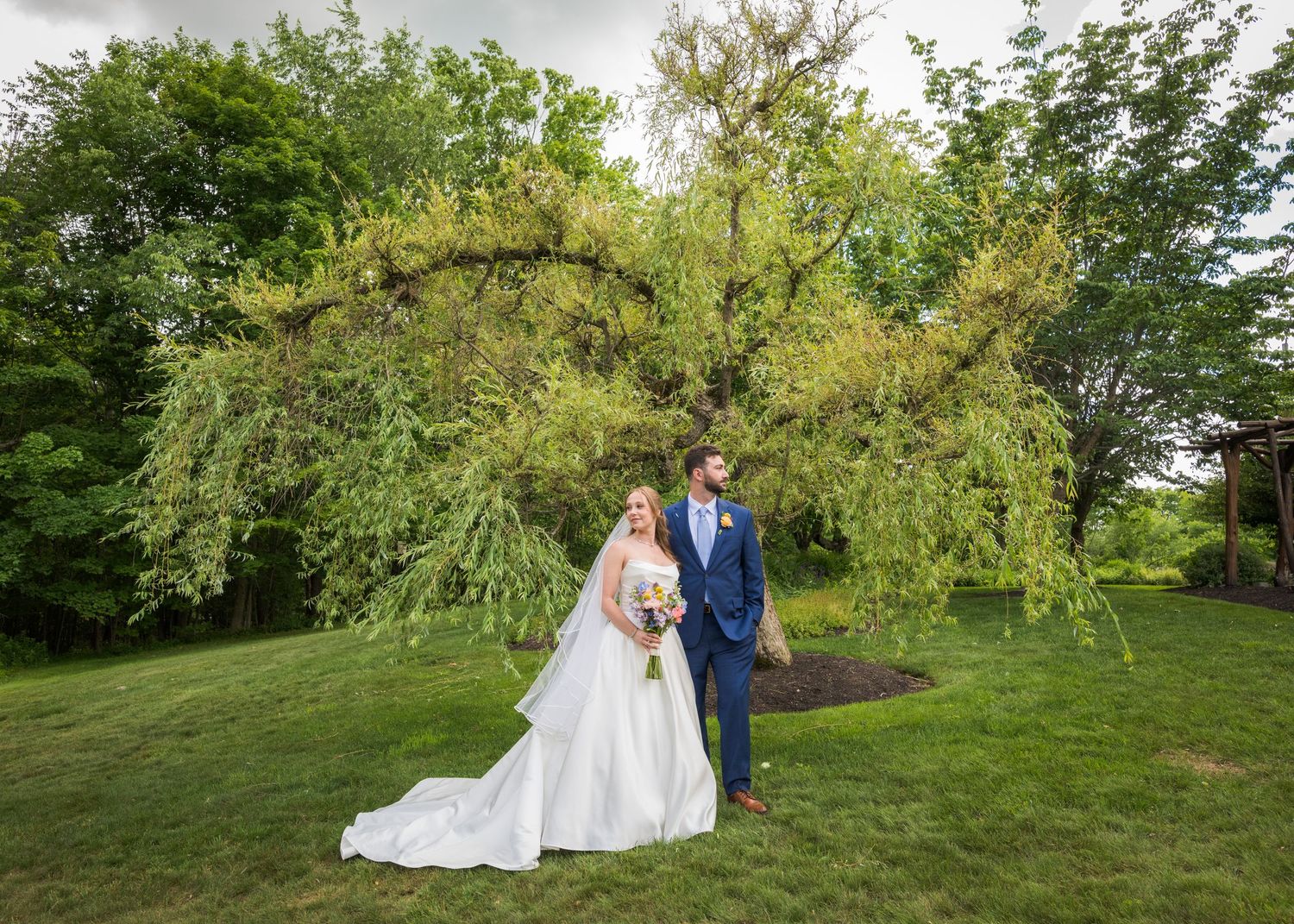 Bride and groom standing together under a sweeping willow tree on lush green lawn at outdoor wedding venue.