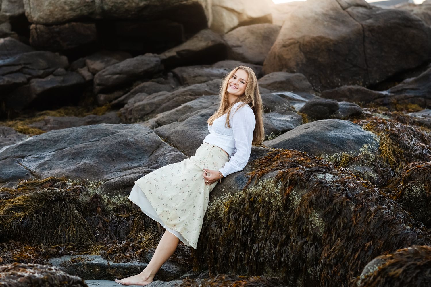 Blond senior girl wears white blouse and yellow floral skirt posing leaning on dark coastal rocks for senior photos in Gloucester, MA.