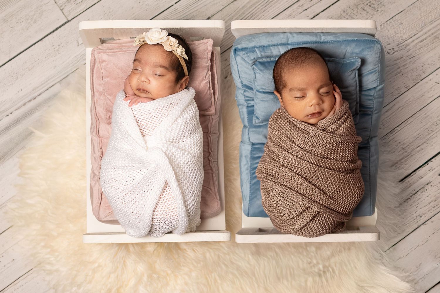 Two swaddled newborn twins sleeping peacefully in small wooden crates on a fluffy white rug.