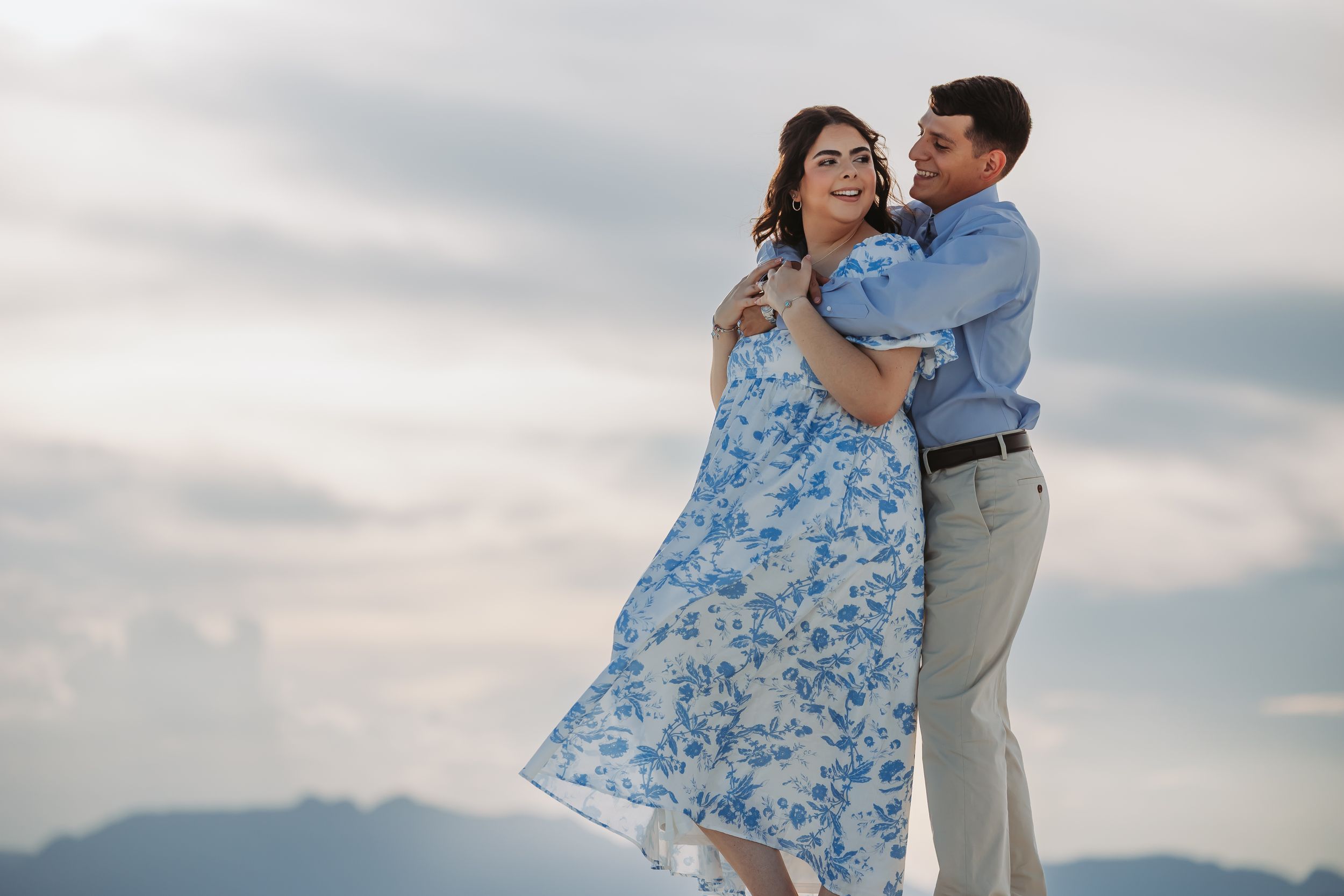 Couple embracing outdoors in blue floral dress and blue shirt with mountains and cloudy sky backdrop.