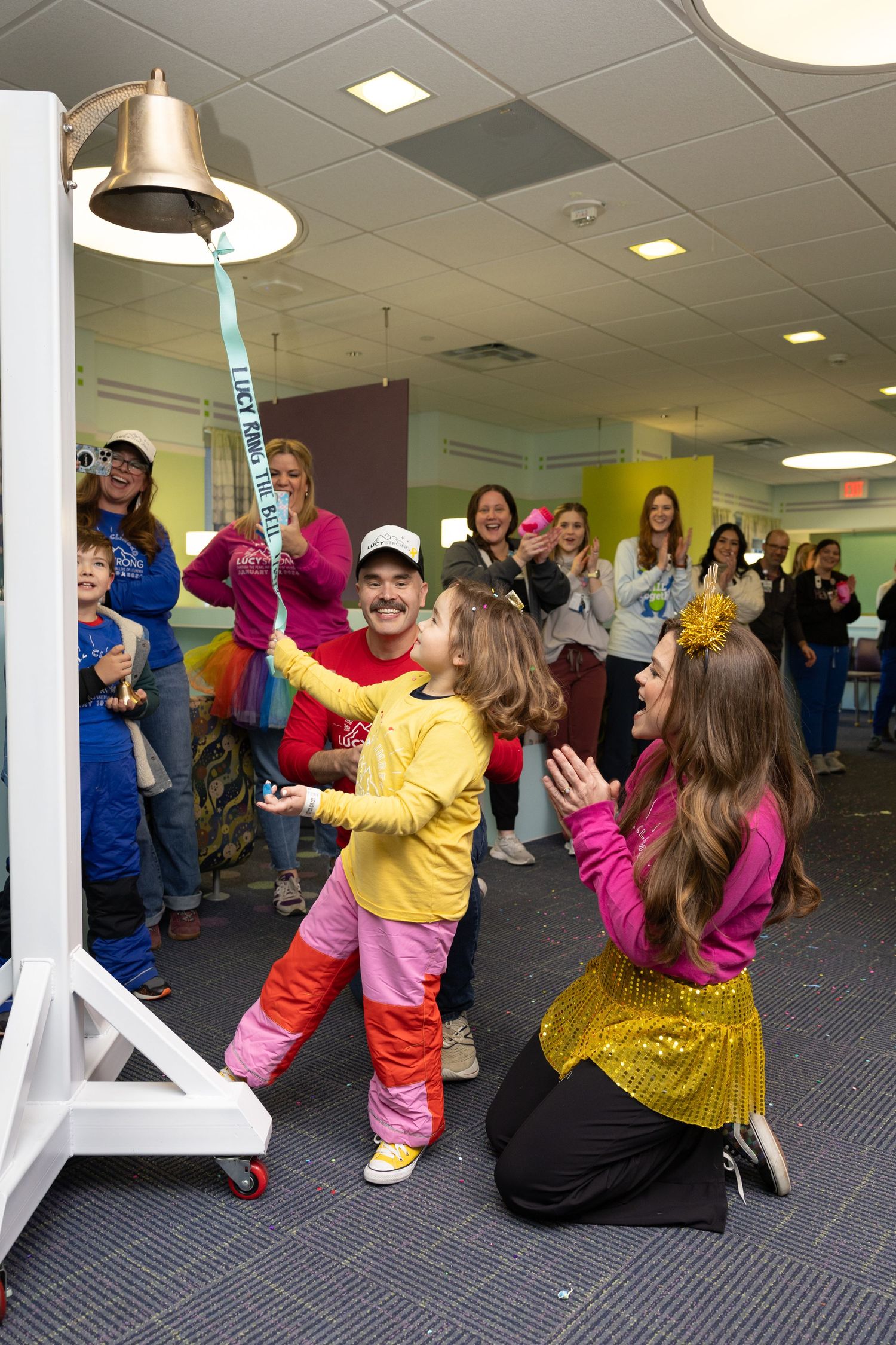 Child in yellow outfit rings a victory bell at a pediatric hospital, cheered on by family and staff.