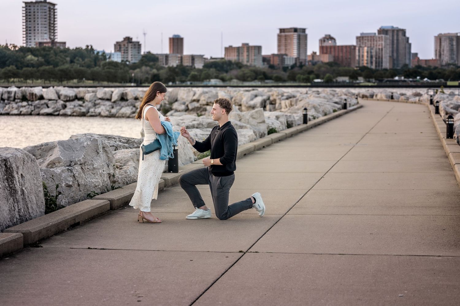 Man proposes to woman on waterfront path with city skyline in background on a romantic evening.