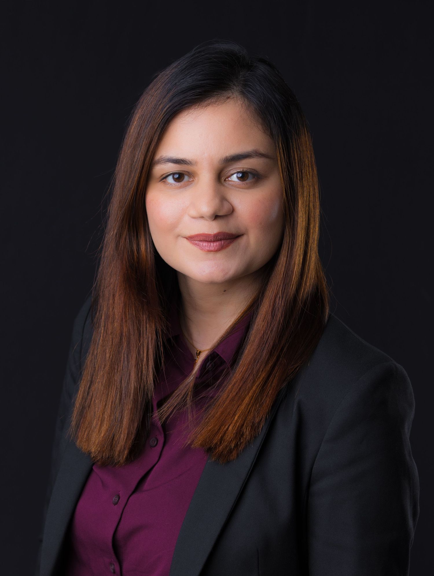 Professional headshot of a woman in a black blazer and maroon shirt with long brown hair against a dark background.
