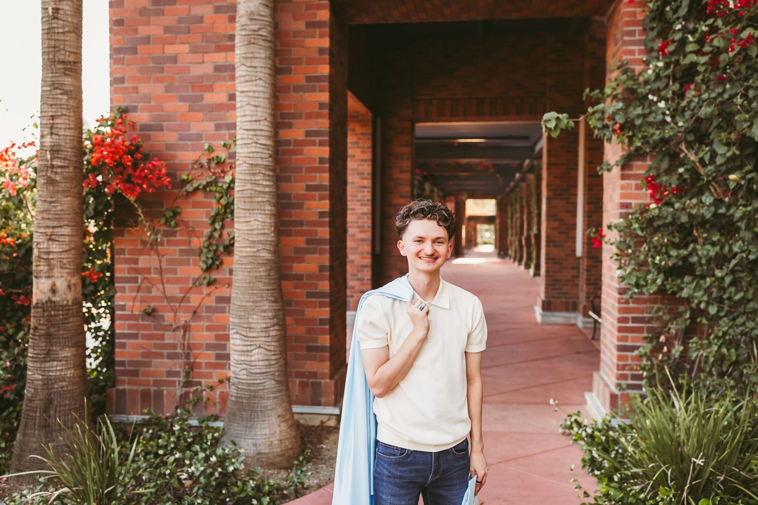 Young man in white polo shirt holding jacket, smiling in a brick archway corridor with red flowers.