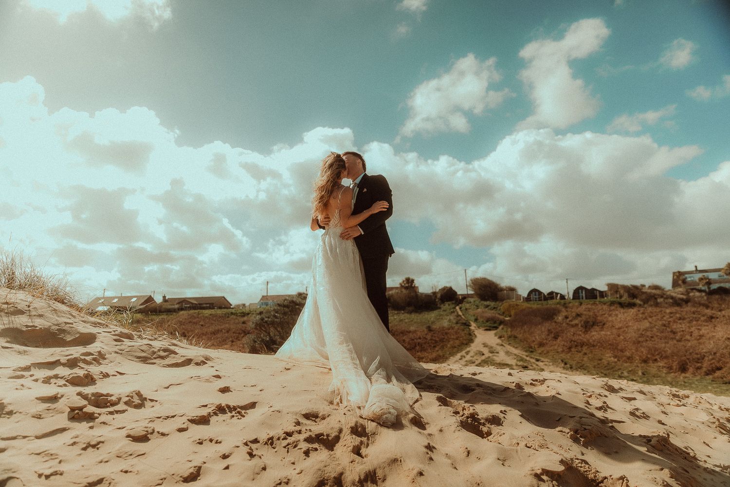 A couple shares a romantic kiss on sandy dunes under a dramatic cloudy sky, bride in flowing white gown.