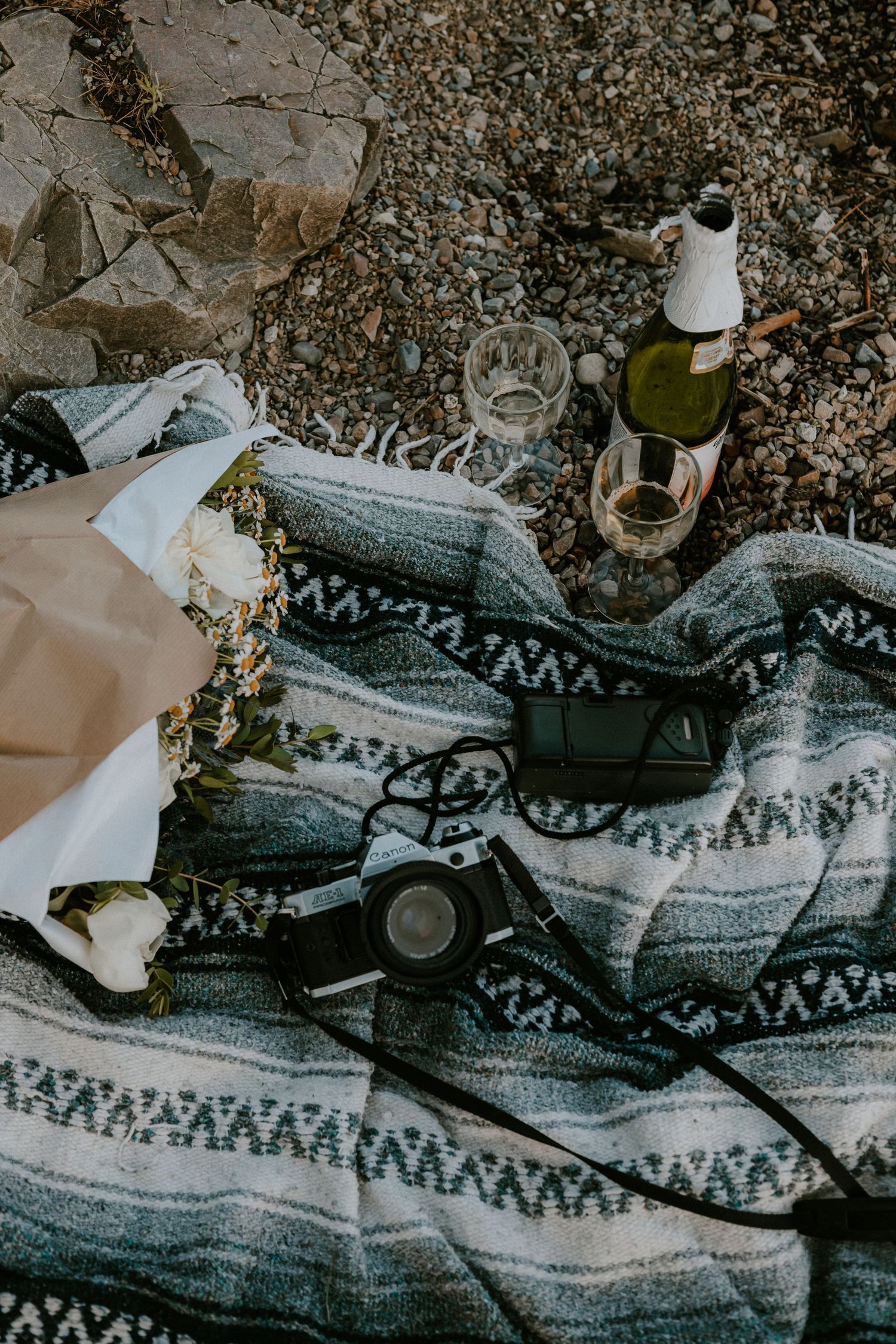 Rustic picnic blanket with wine bottle, glasses, camera, and floral bouquet arranged on rocky ground outdoors.