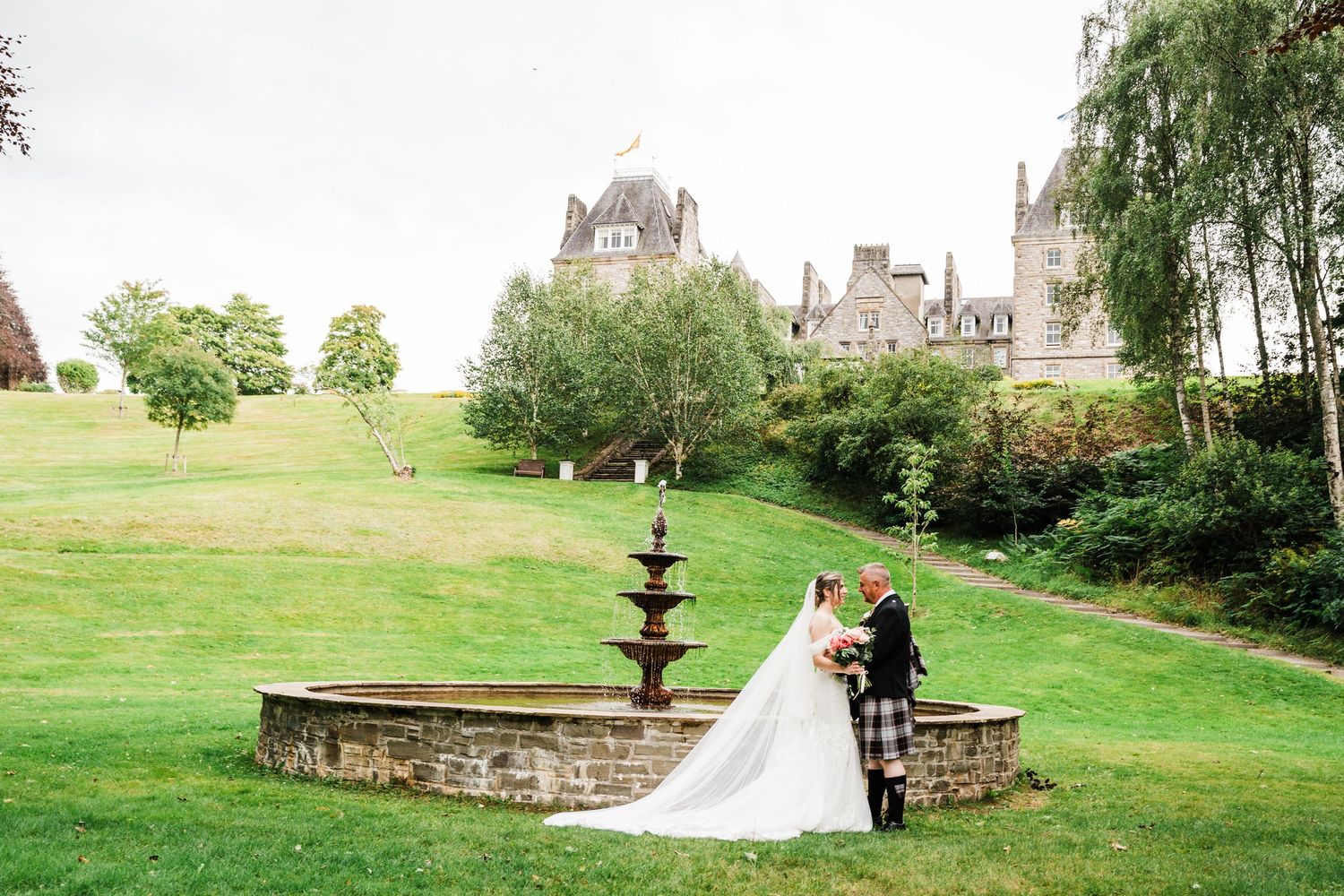 Bride and groom outside Atholl palace in Pitlochry, next to fountain with hotel in background Groom in kilt outfit, bride in white