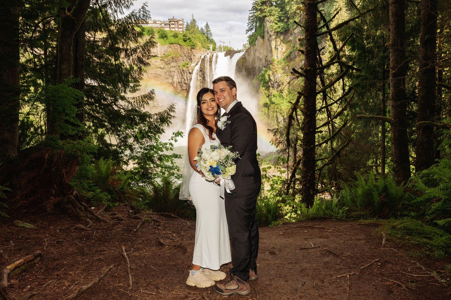 Bride and groom pose in front of stunning Snoqualmie Falls waterfall surrounded by lush green forest on their wedding day.