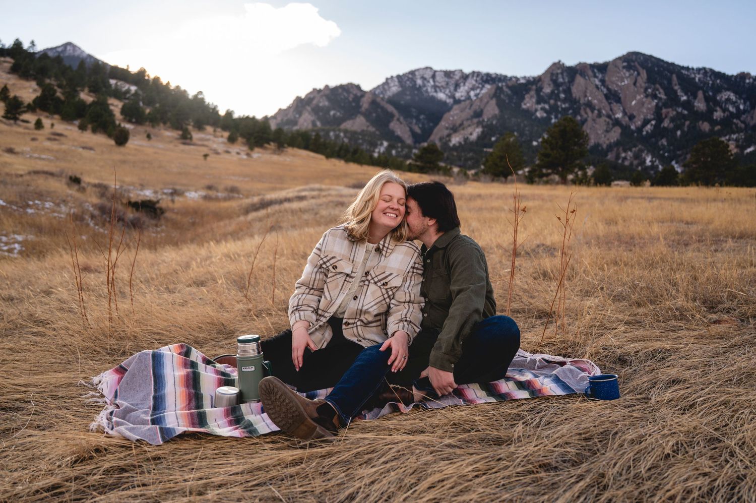 Couple sitting on a blanket in a golden field with Rocky Mountain Flatirons backdrop during a romantic outdoor picnic.