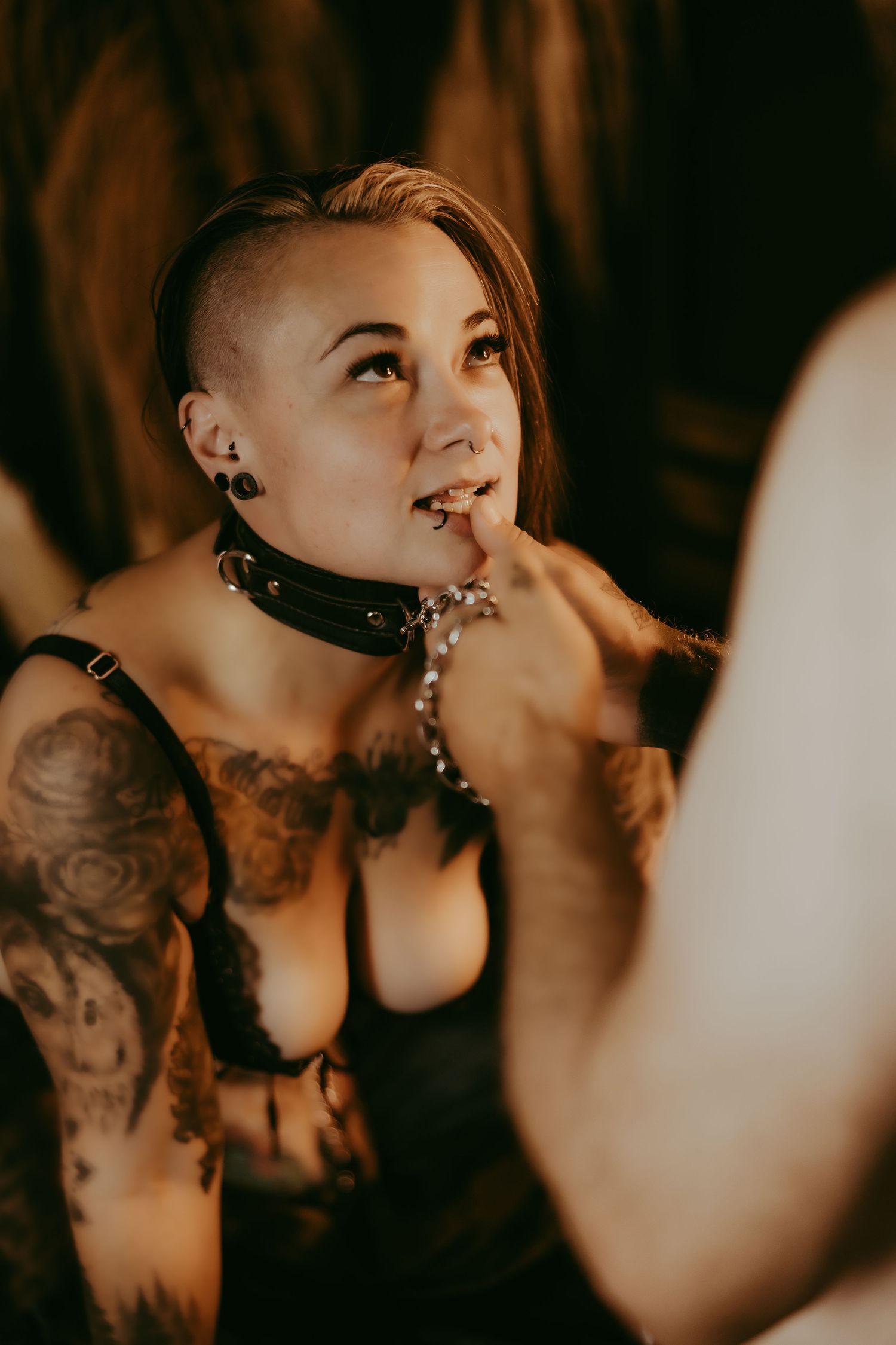 Tattooed woman with shaved sides, leather collar, and black lingerie posing indoors with a thoughtful expression.