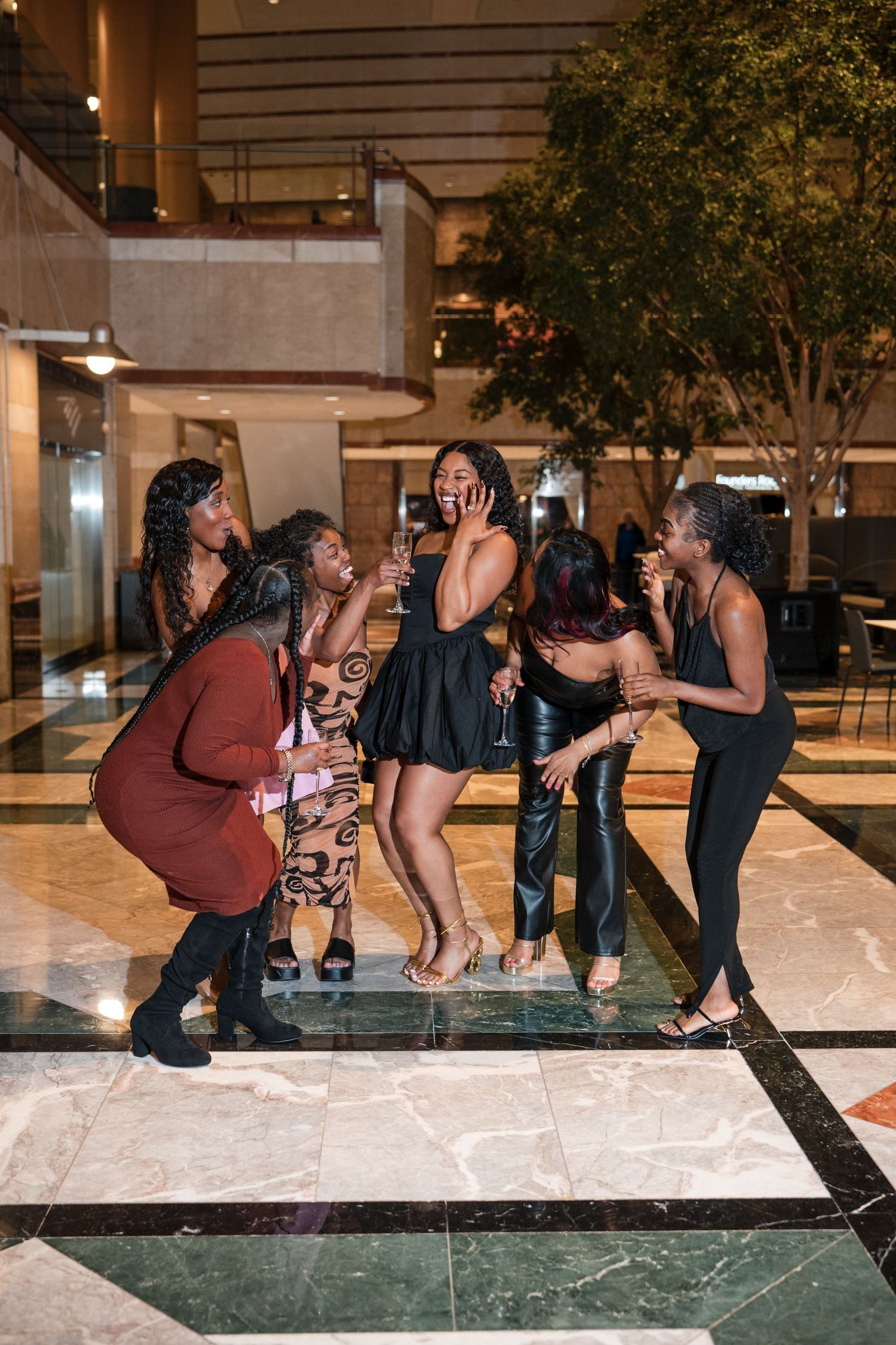 Group of six women laughing and celebrating together in an elegant marble lobby during a night out.