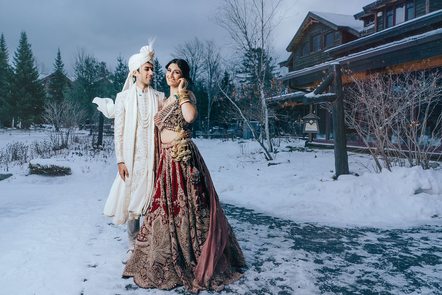 Bride and groom in traditional Indian wedding attire standing in the snow outside a rustic lodge in Lake Placid, NY