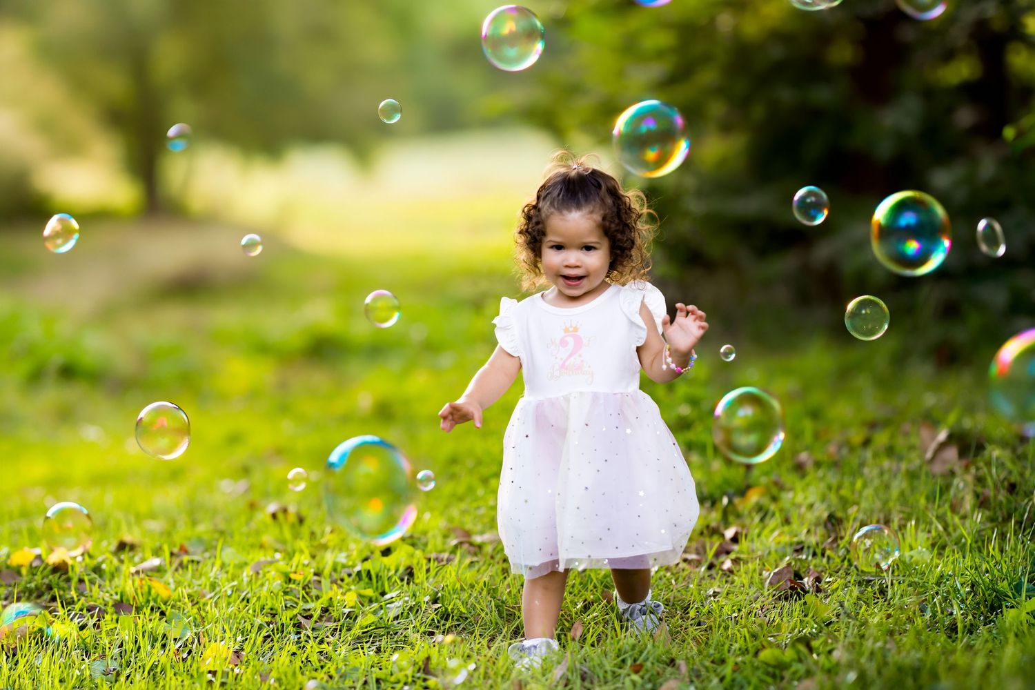 A joyful toddler girl in a white dress plays with colorful soap bubbles on a sunny green lawn.