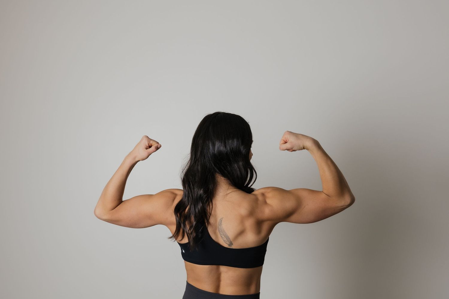 Muscular woman flexing both arms showing strong back muscles, wearing black sports bra, rear view on white background.