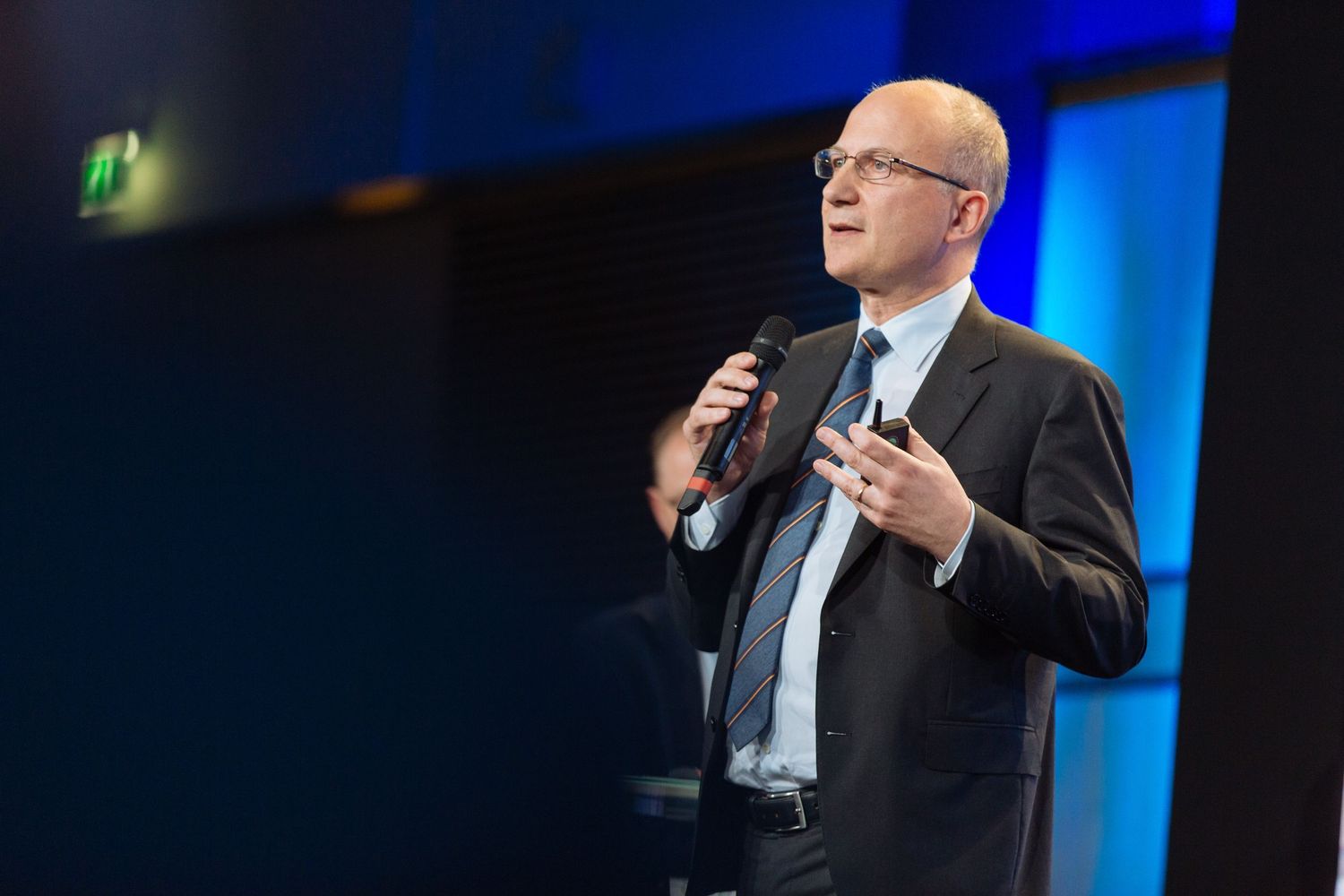 Professional male speaker holding microphone on stage with blue lighting at a corporate conference event.