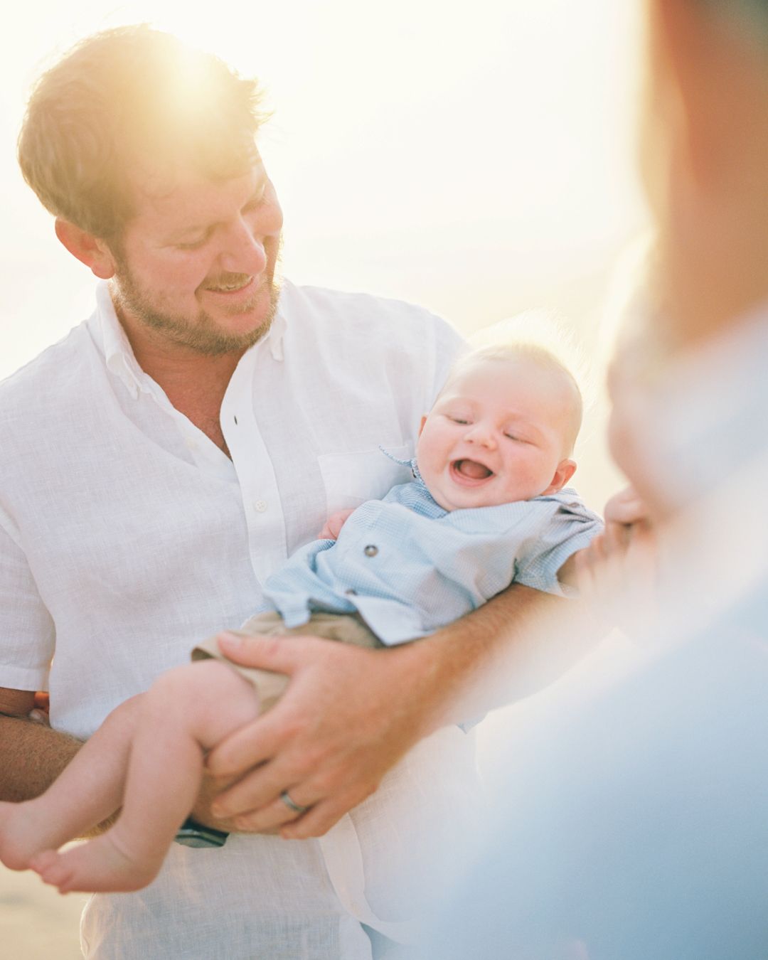 A parent in white clothing holds and plays with a laughing baby wearing light blue in a bright sunlit setting.