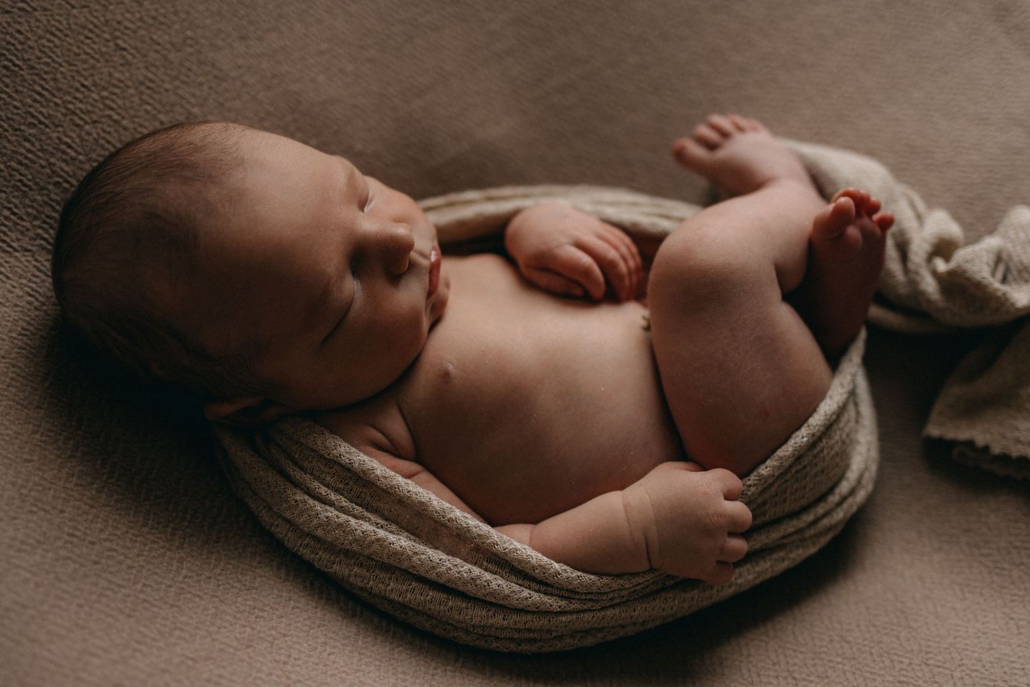 Sleeping newborn curled up on brown fabric in artistic pose.