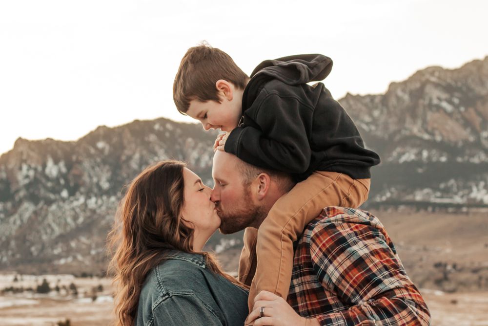 A couple kisses while their young son sits on dad's shoulders against a scenic mountain backdrop | How I Help You Prep for Family Photos