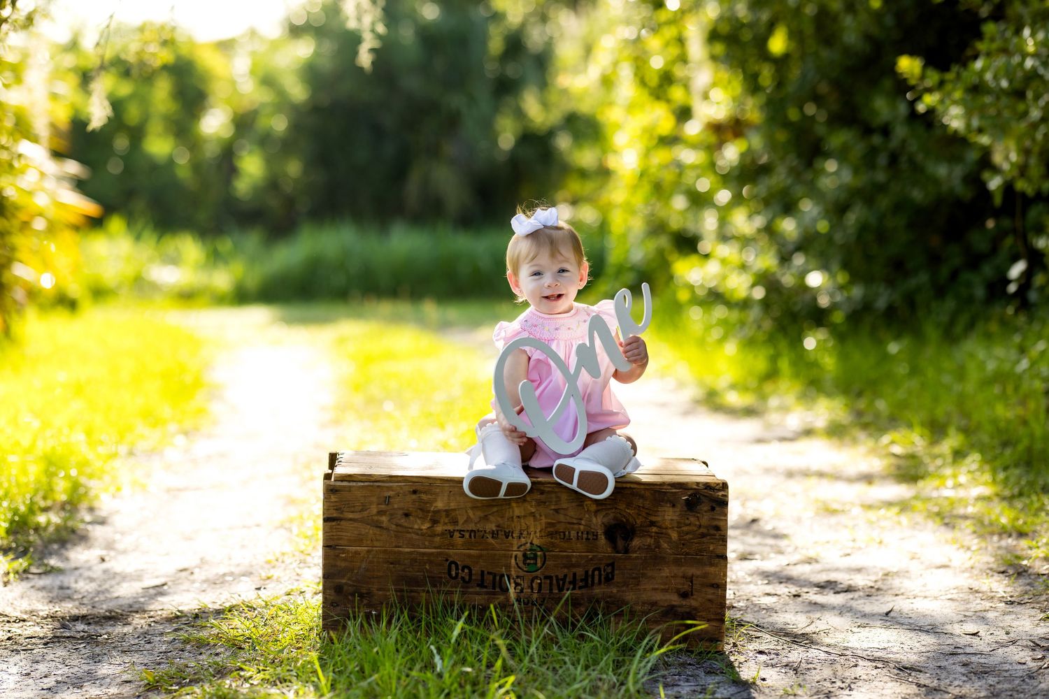 Smiling baby girl in pink outfit sitting on rustic wooden crate outdoors in sunny green park setting.