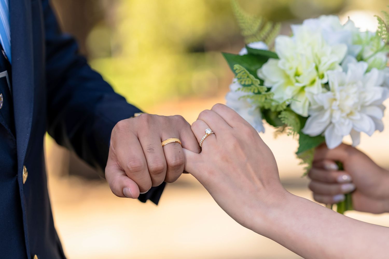 Newlyweds holding hands with wedding rings, bride holding white dahlia bouquet at outdoor ceremony.
