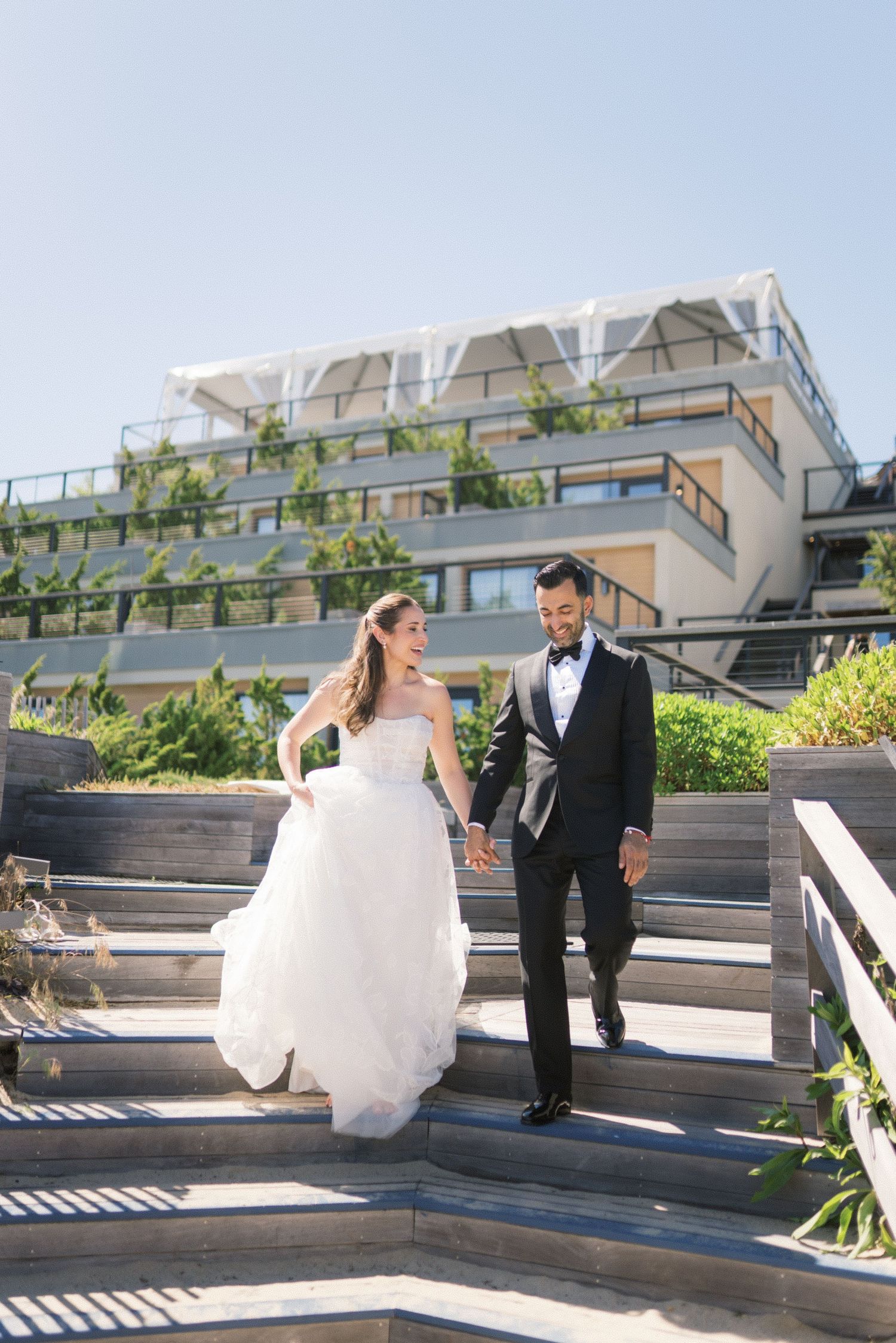 Smiling bride in white lace gown holding vibrant pink and coral bouquet with peonies and anthuriums outdoors.
