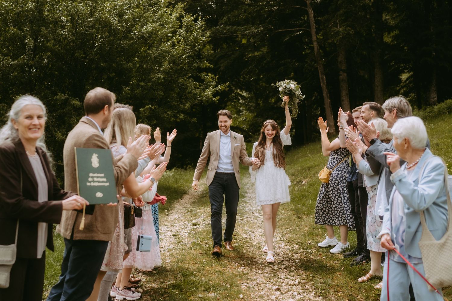 Newlywed couple walks down a forest path while guests clap and celebrate during an outdoor wedding ceremony.