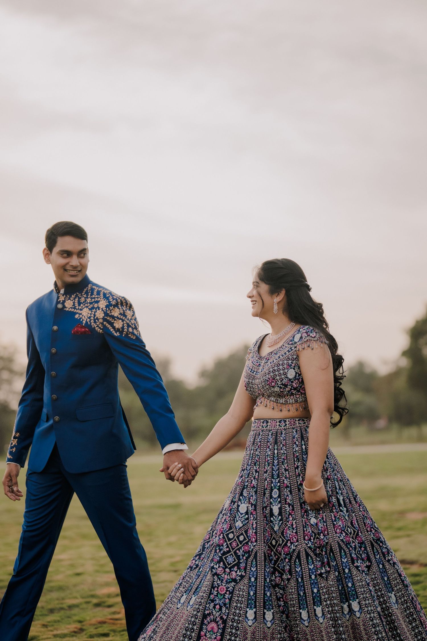 Couple in traditional Indian wedding attire holding hands outdoors, groom in blue sherwani, bride in embroidered lehenga.