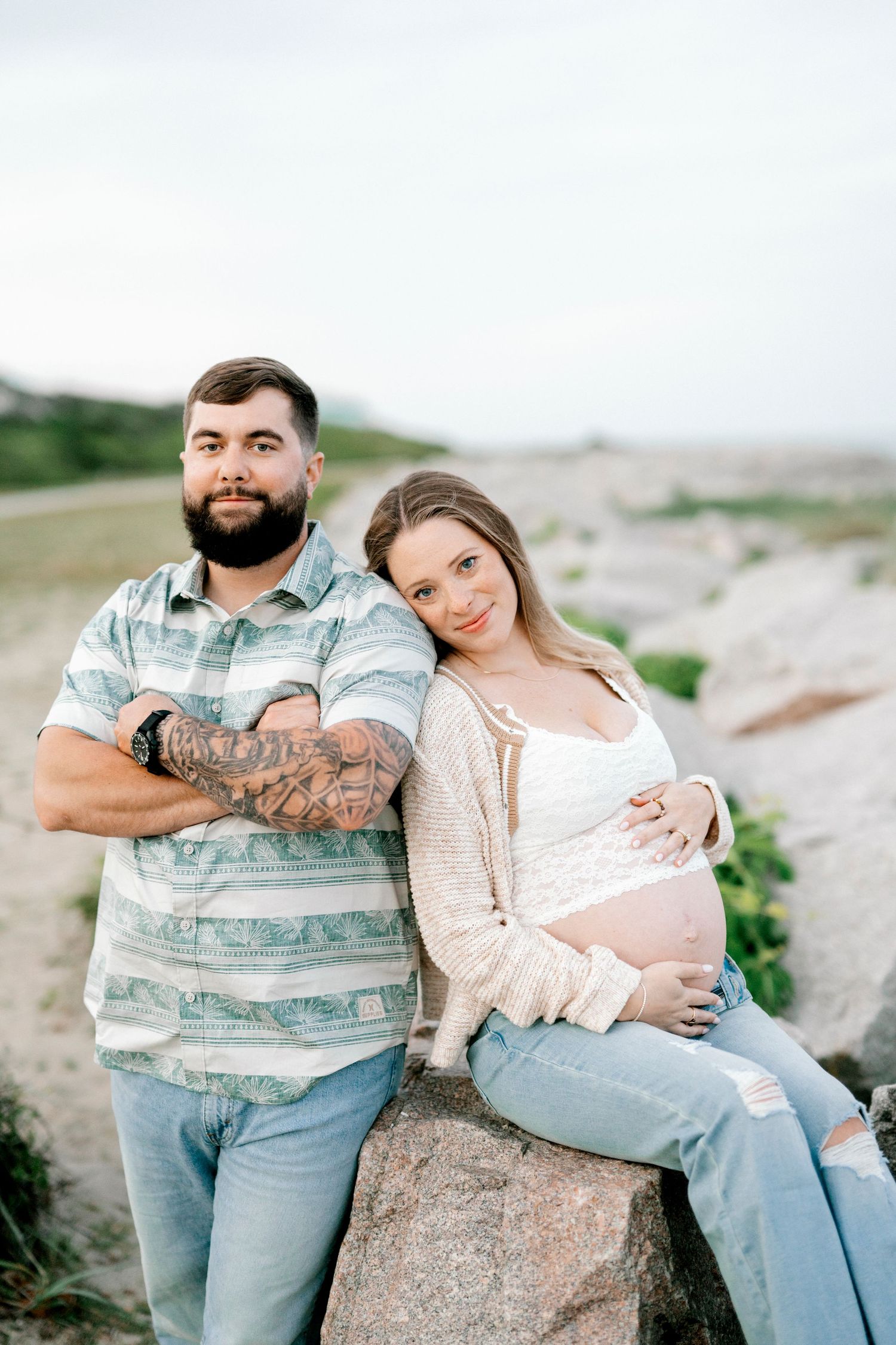 A bearded tattooed man and pregnant woman pose together on rocks outdoors in a maternity photo shoot.