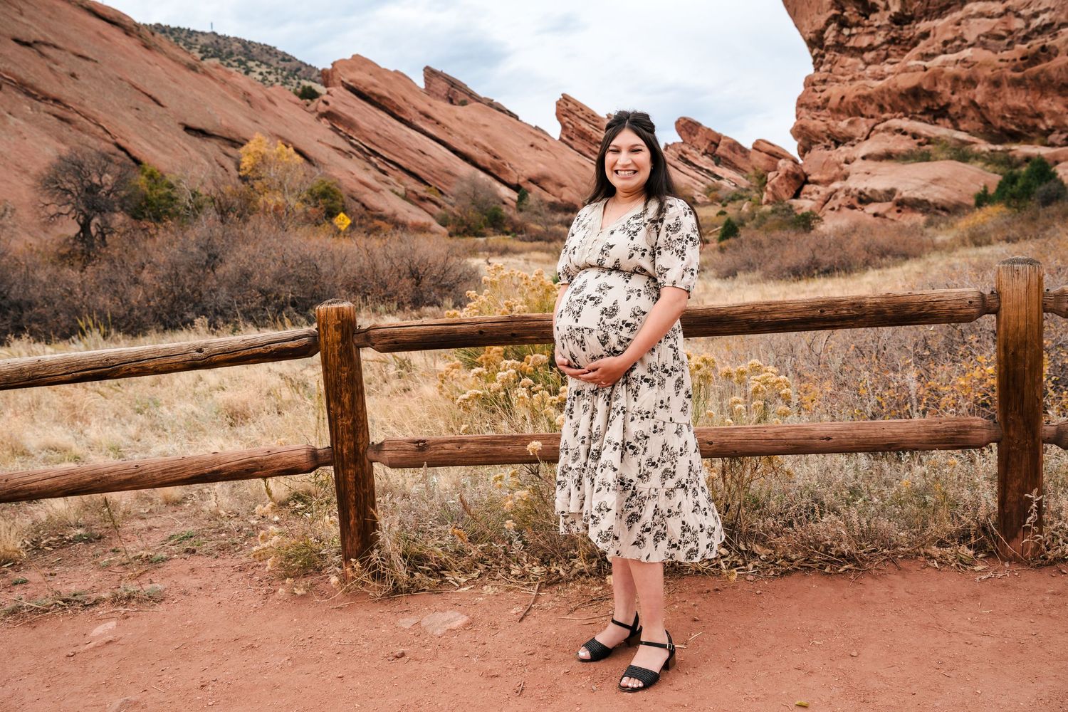 Pregnant woman in floral dress posing by wooden fence at Red Rocks Park with red rock formations behind her.