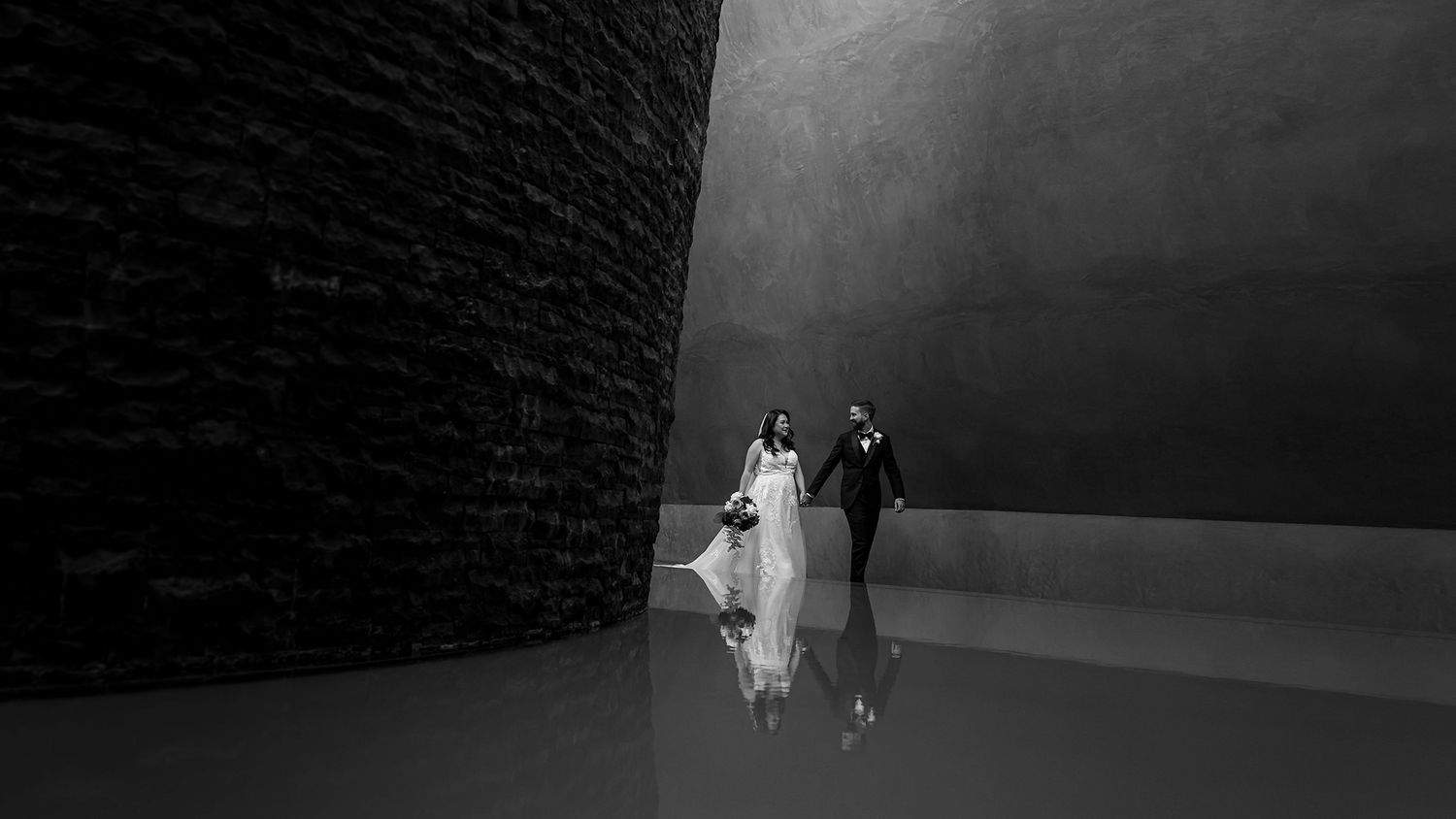 Black and white photo of a bride and groom walking hand in hand through a dramatic modern architectural space.