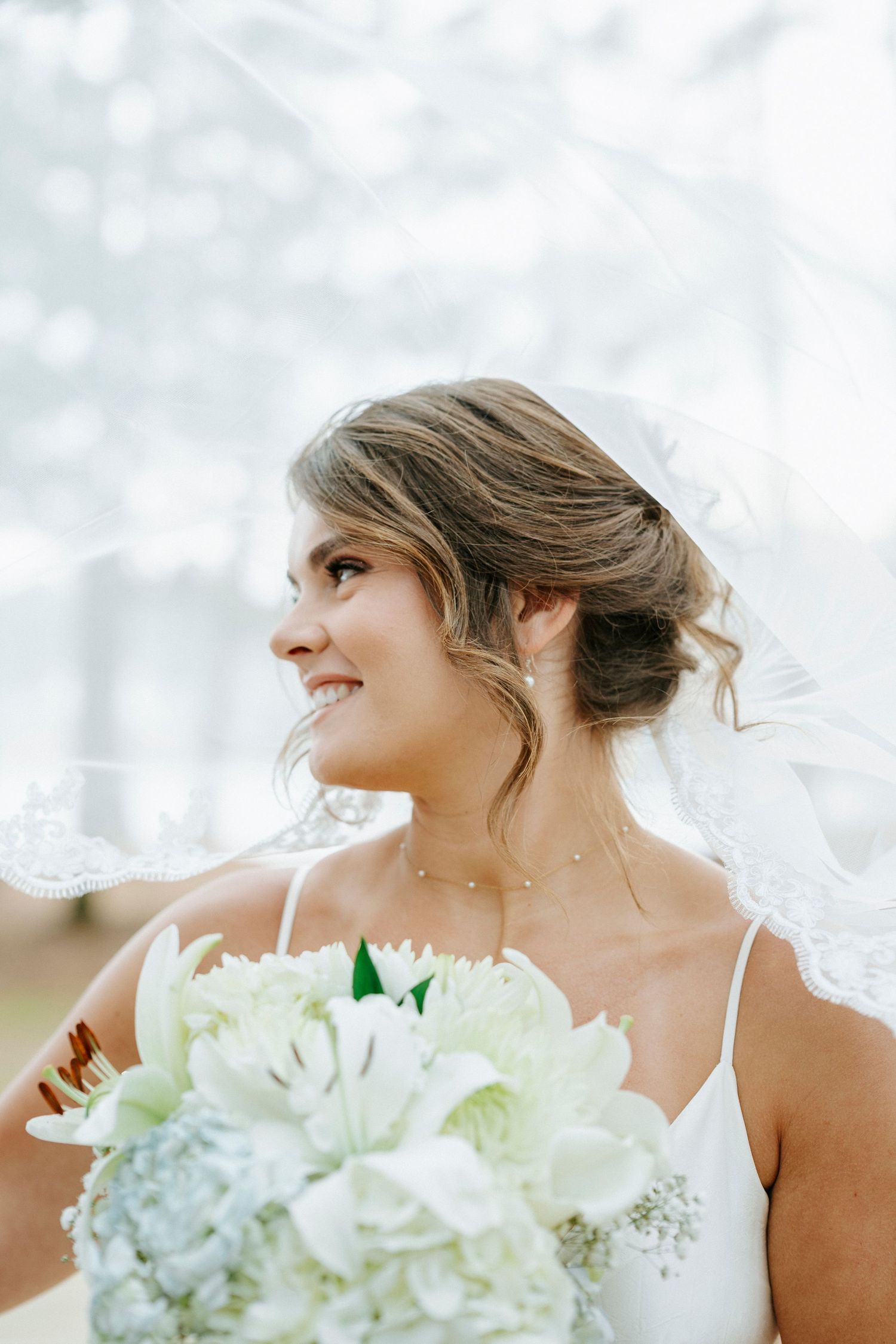 A bride in a white dress holds a white floral bouquet while looking to the side with a joyful expression.