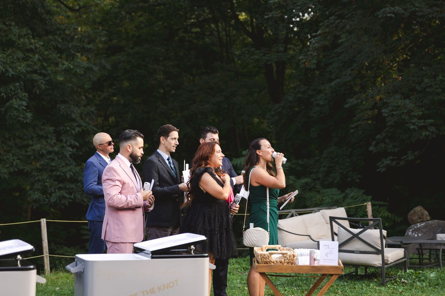 Wedding guests in colorful garden attire enjoy drinks from cooler amidst greenery and comfortable lawn furniture at a Hudson Valley wedding.