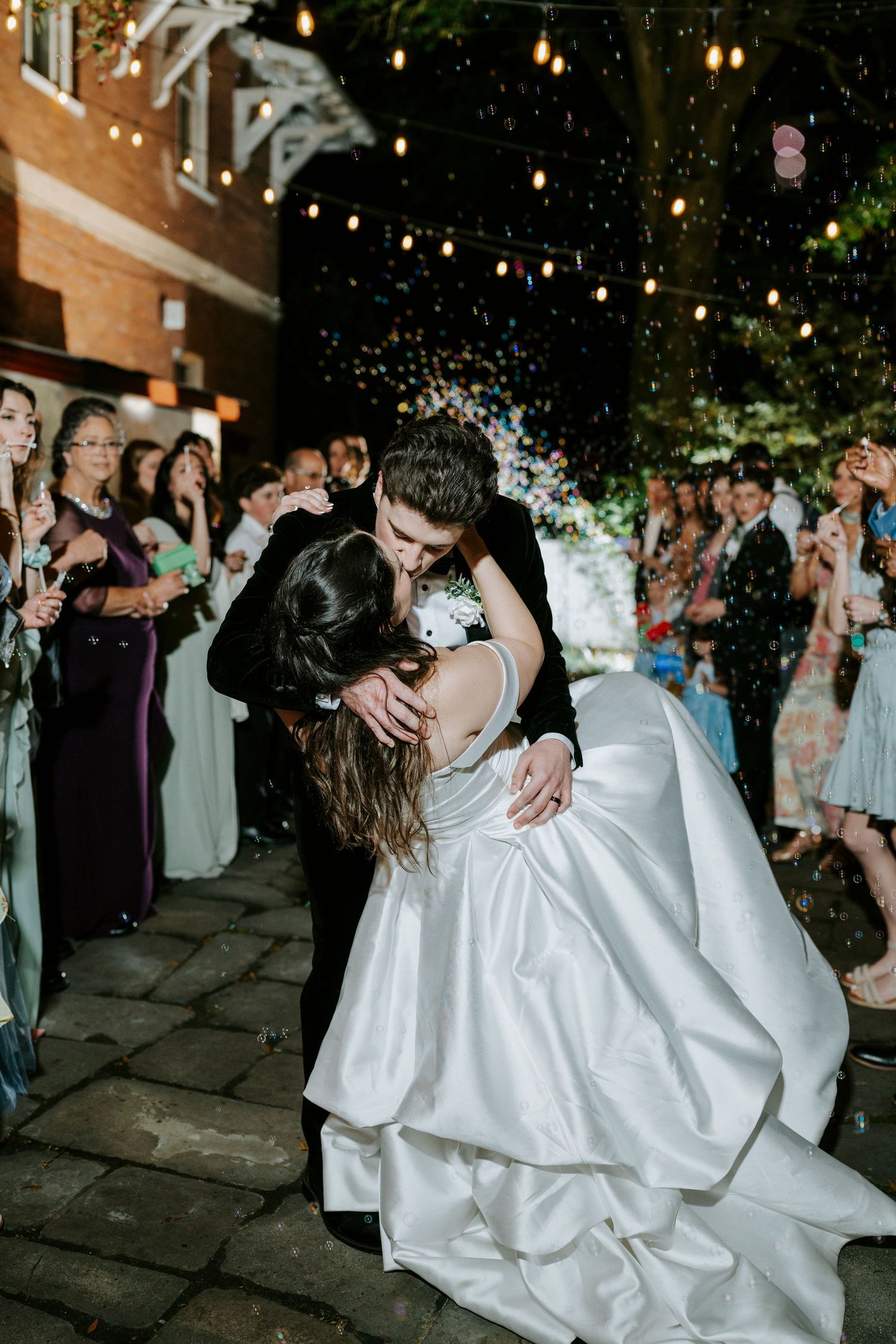 A couple shares their first dance as newlyweds under romantic string lights while guests look on at an outdoor evening reception.