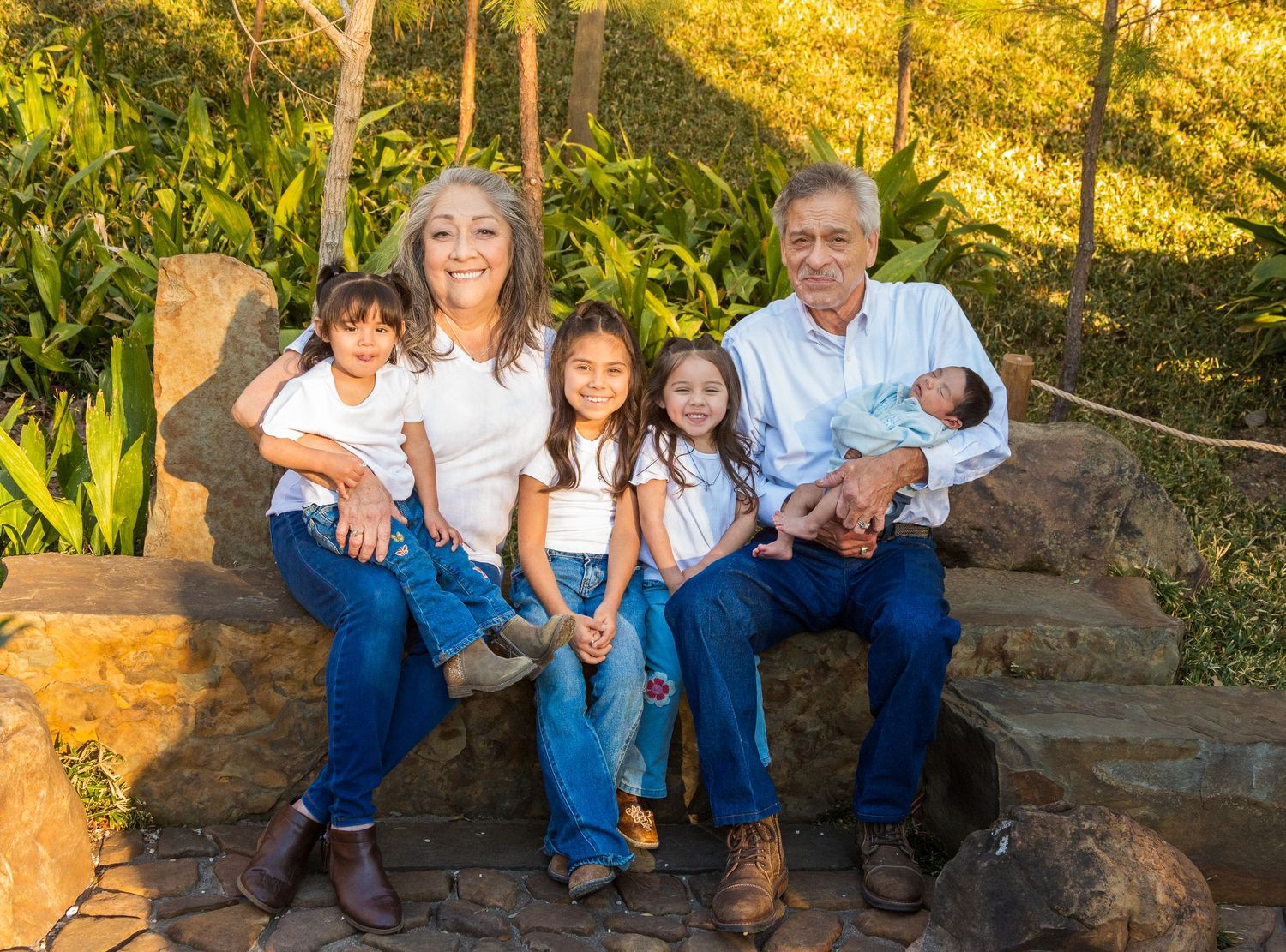 Grandparents sitting outdoors on stone steps with four young grandchildren, smiling in a sunny garden setting.