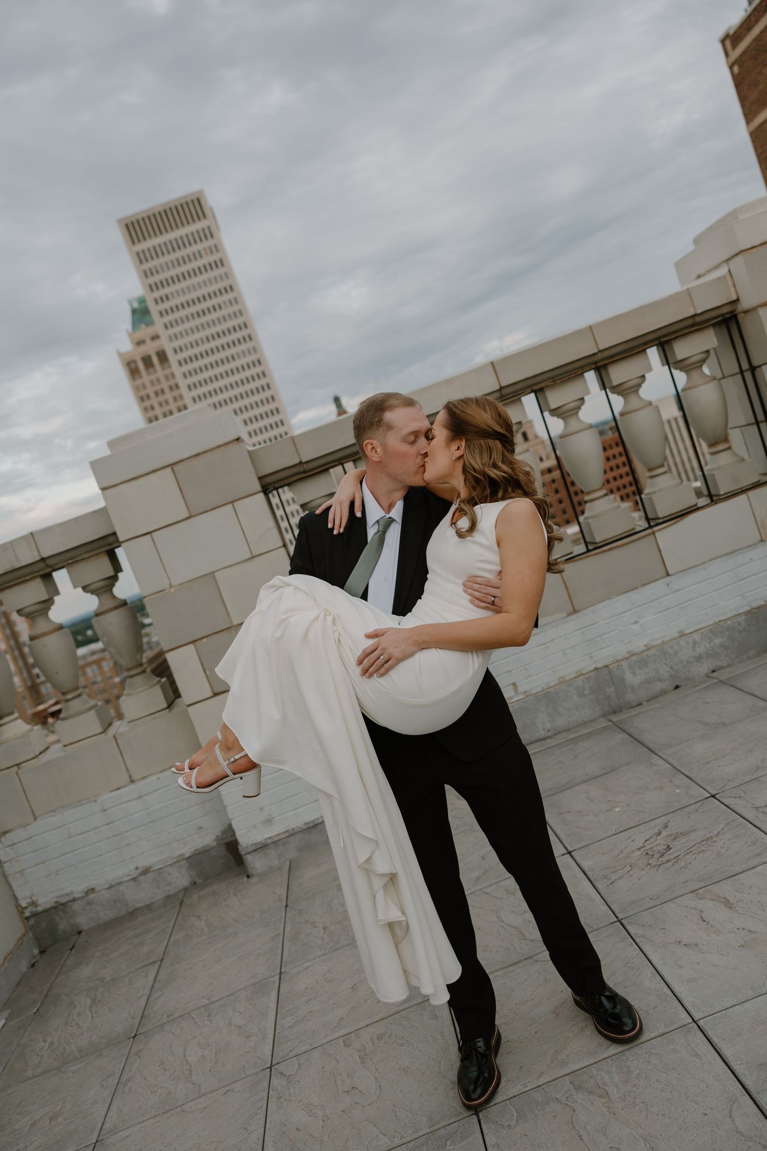 Groom carries bride in white dress on rooftop terrace, sharing a kiss with city skyline in background.