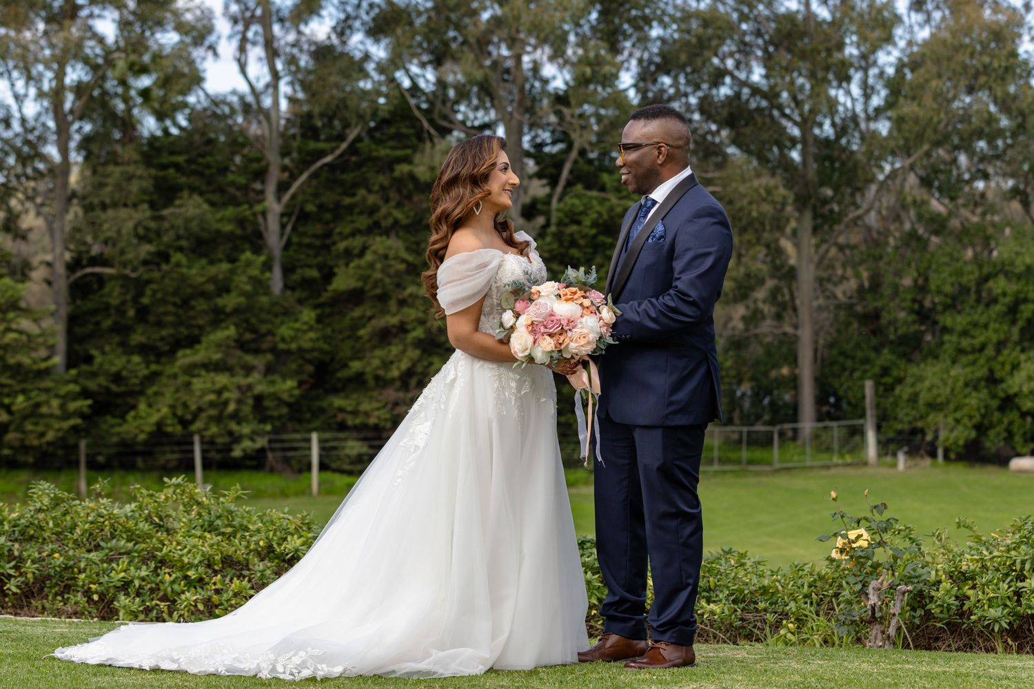 Bride and groom stand facing each other in lush garden, she in white off-shoulder gown holding pink bouquet.