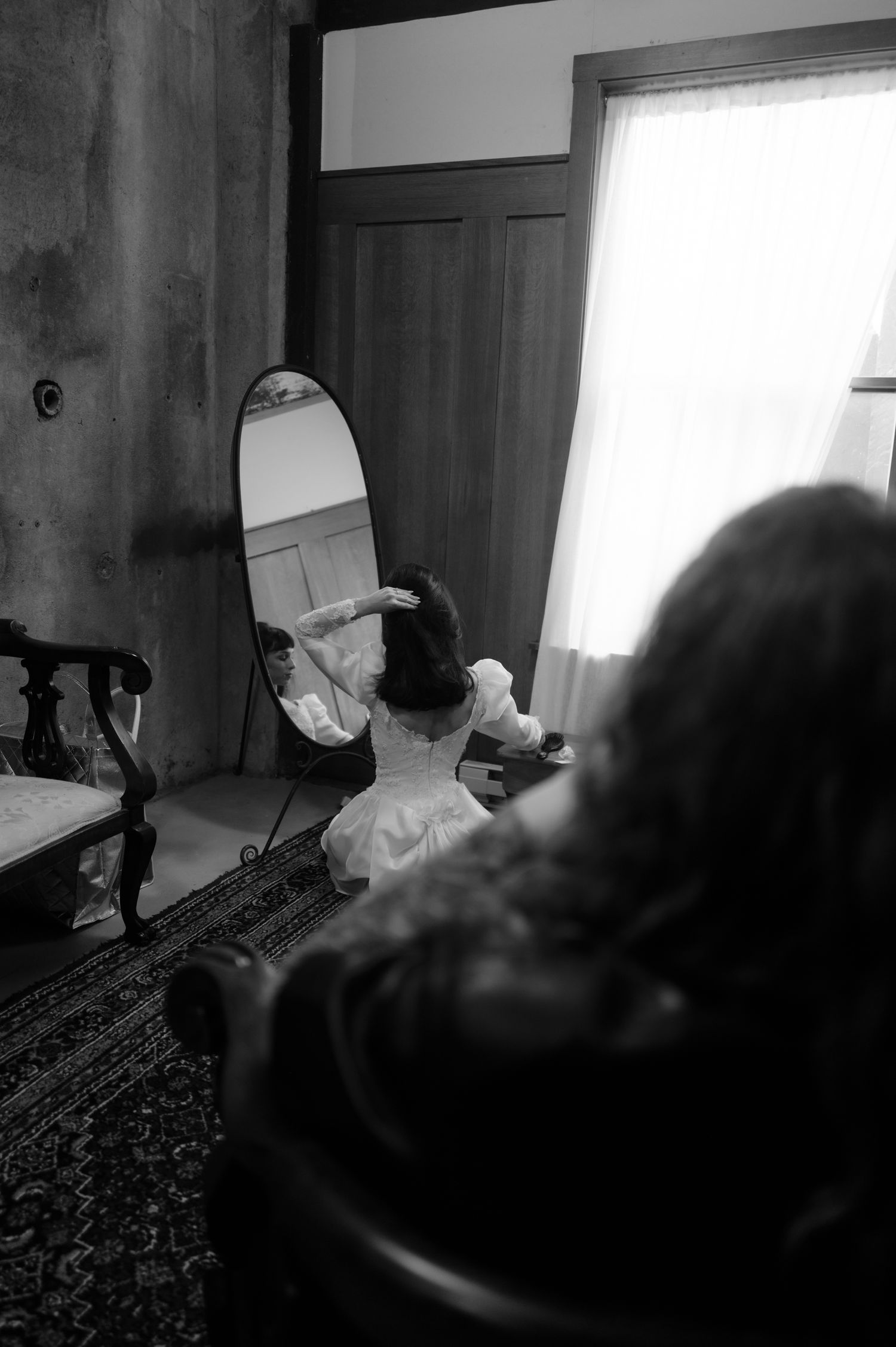 Black and white photo of a bride in white dress adjusting hair while seated before an oval mirror near a window.