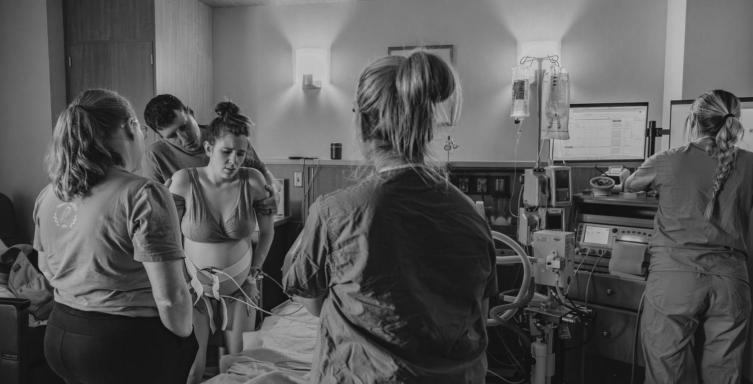 Two adults lovingly kiss a newborn baby in a hospital room while golden sunlight streams through the window.
