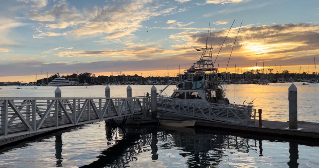 A panoramic view of a marina at sunset with boats silhouetted against a colorful sky and reflective water.