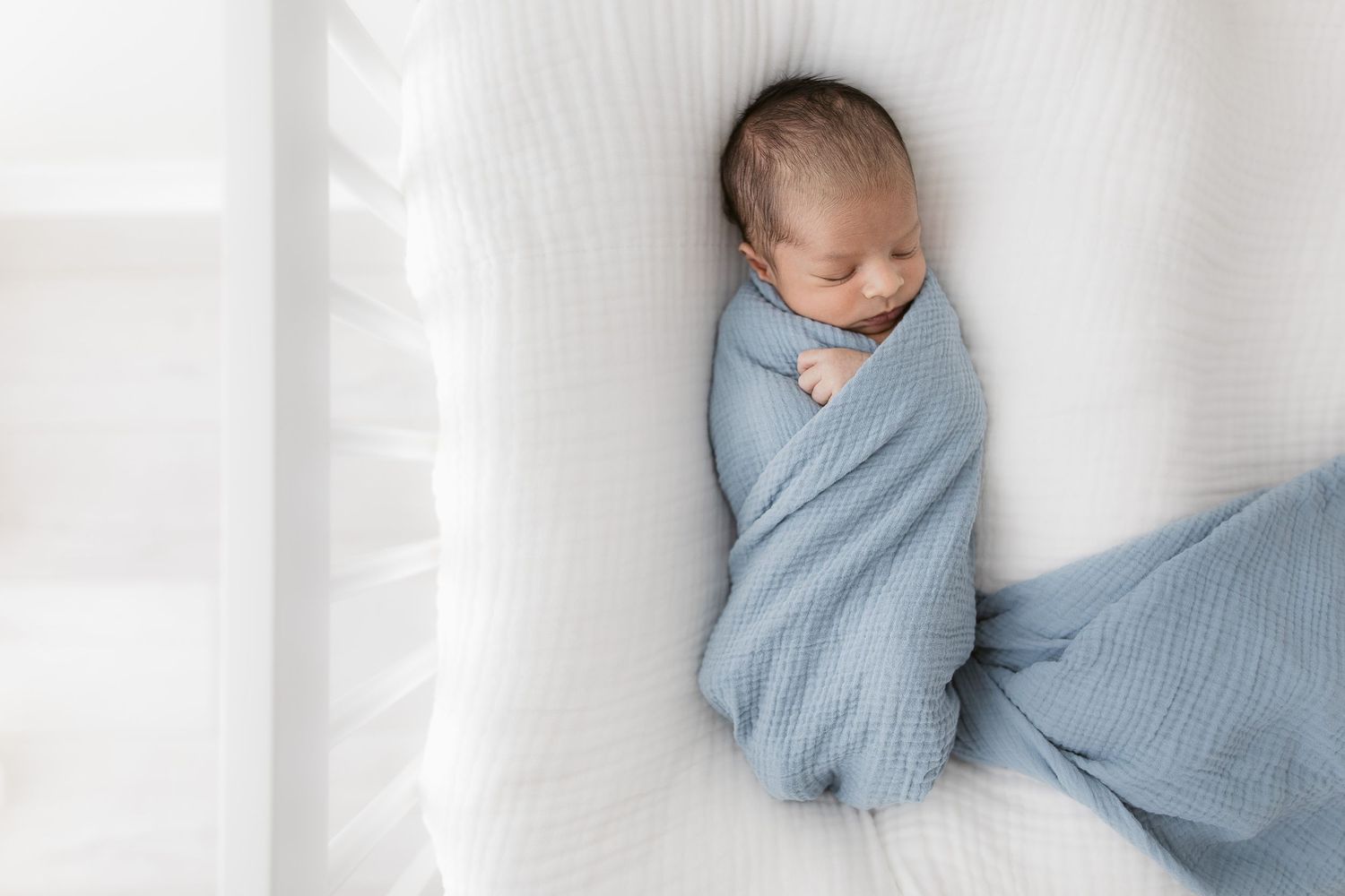 A sleeping baby swaddled in soft blue blankets lies on a white textured surface in natural light.