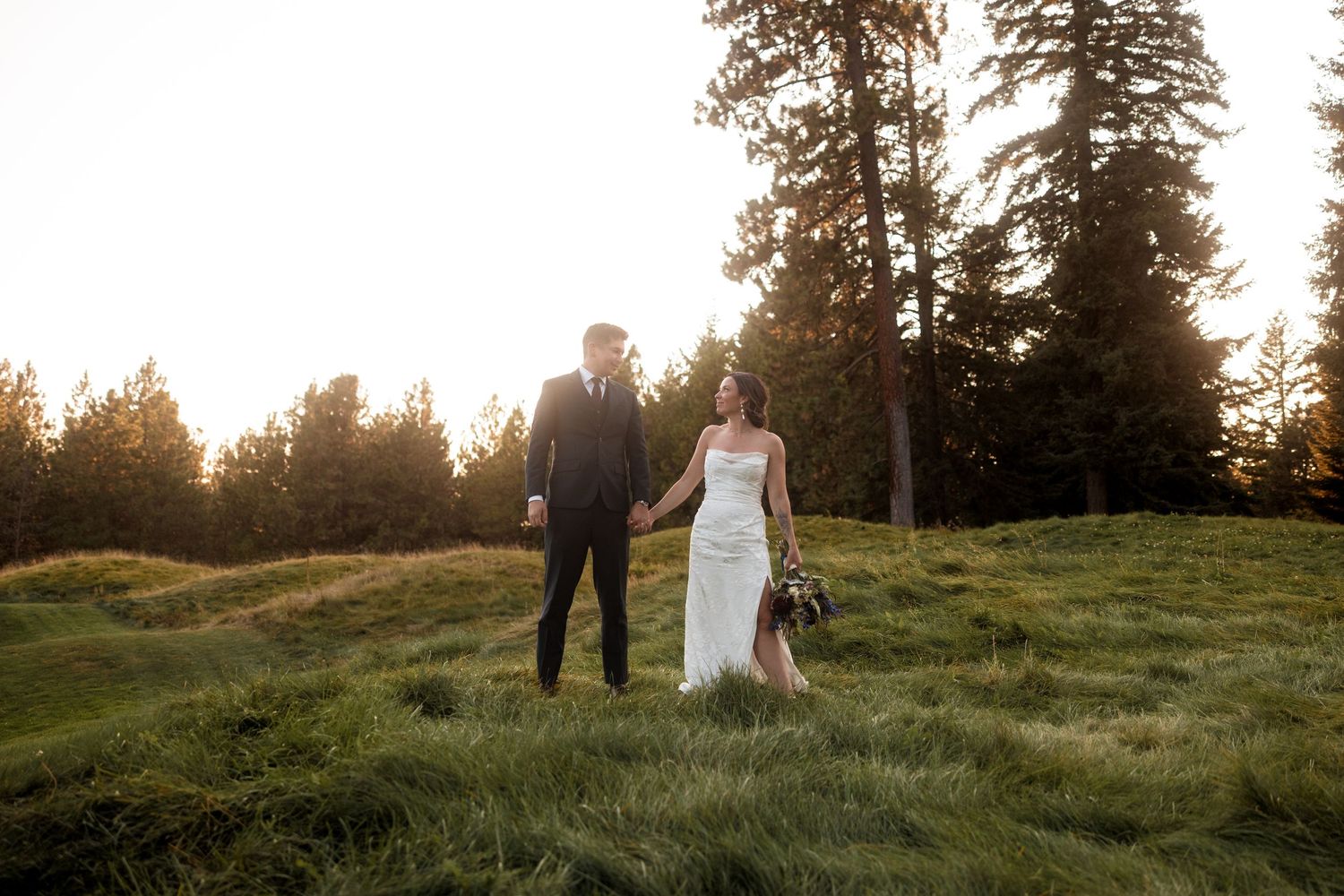 A couple walks hand in hand through a scenic mountain meadow at sunset with tall evergreen trees in the background.