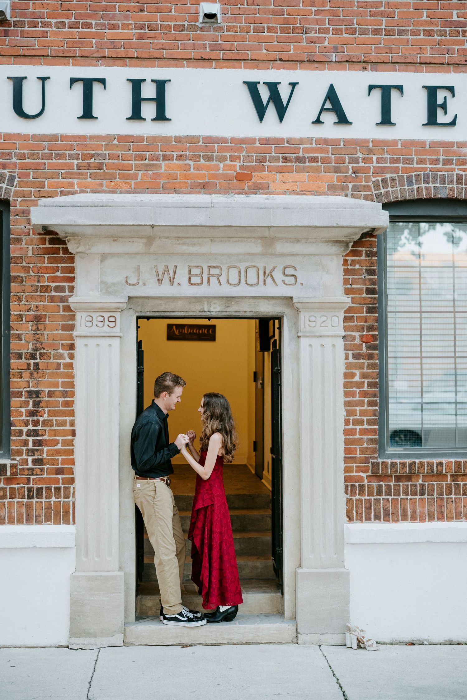 Couple shares a romantic moment in front of a brick building entrance with white stone details and signage.
