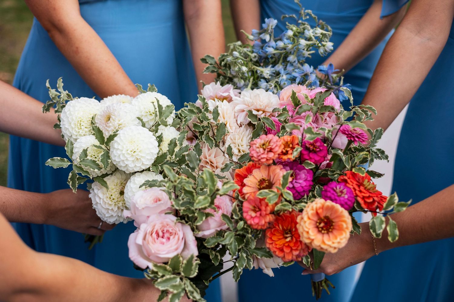 Bridesmaids in blue dresses hold vibrant bouquets featuring white peonies, pink roses, and coral-colored flowers.
